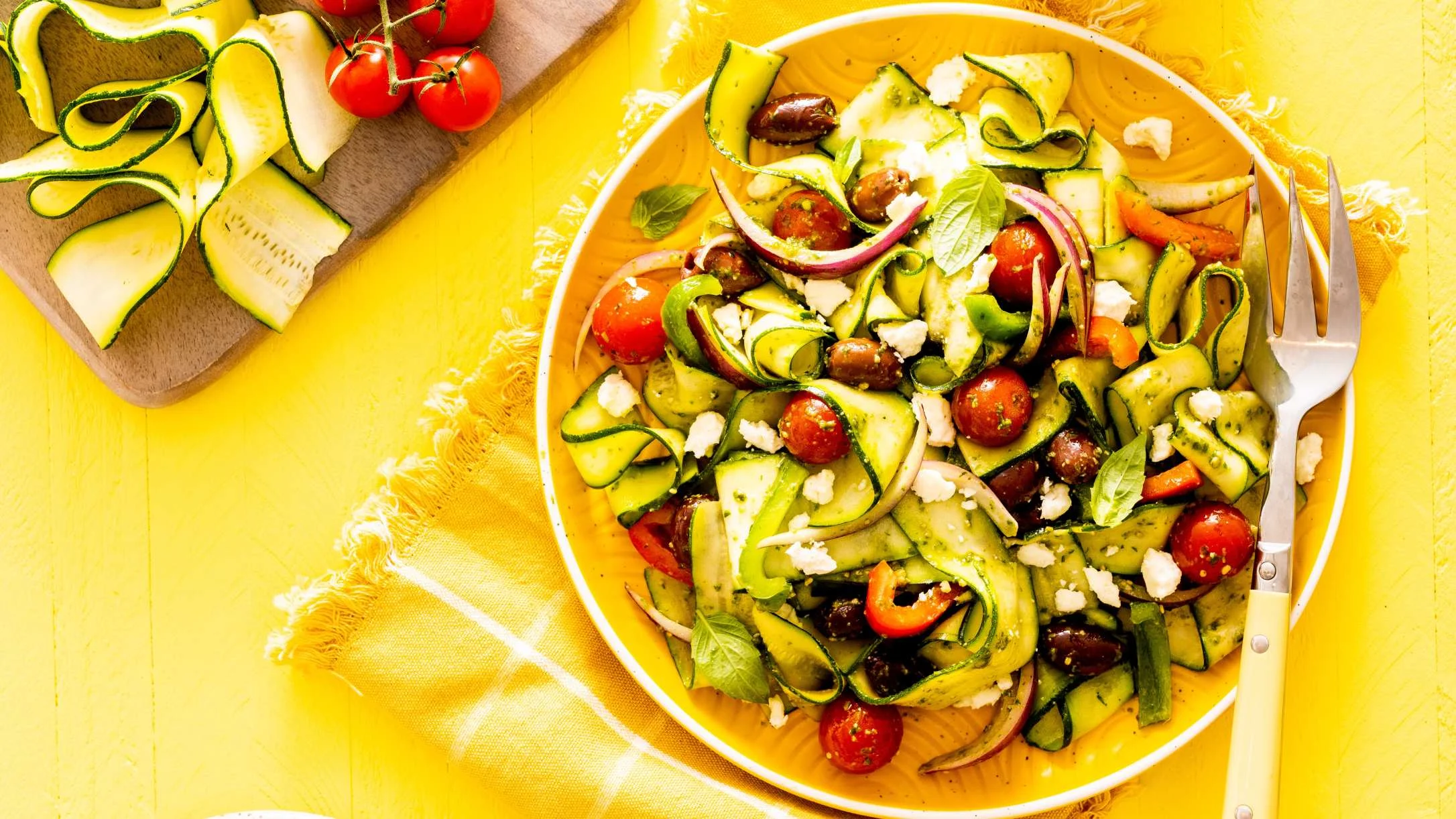 Fresh Mediterranean salad with zucchini ribbons, cherry tomatoes, olives, and feta cheese on a bright yellow background.