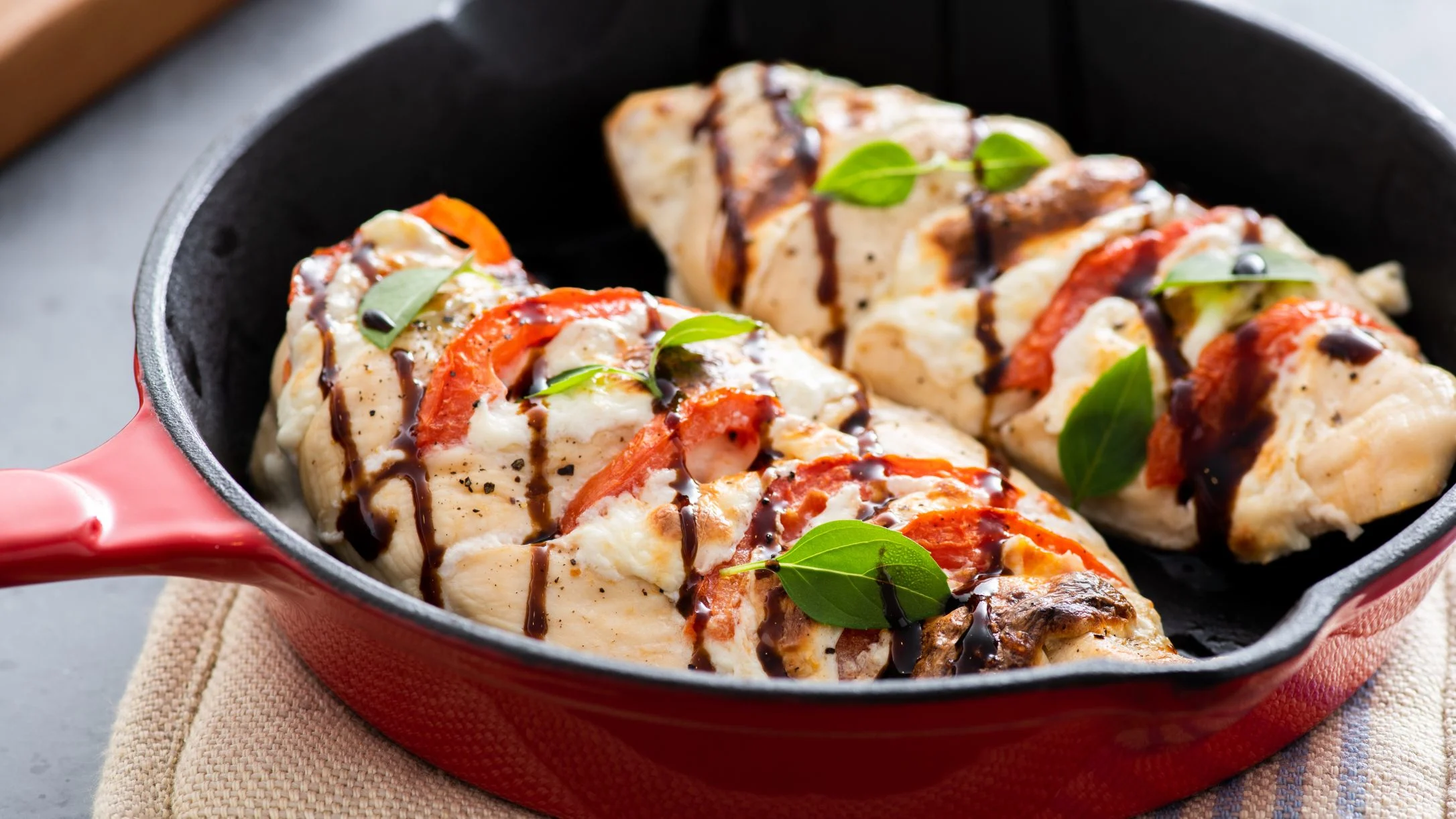 Grilled chicken breasts topped with melted cheese, tomatoes, balsamic glaze, and fresh basil in a red cast iron pan.
