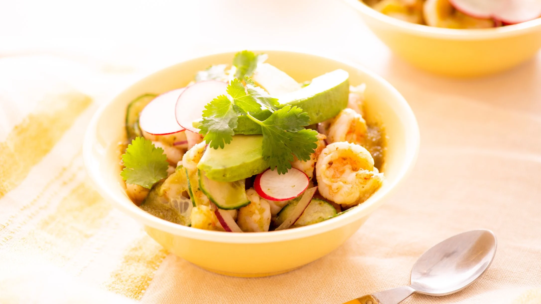 A yellow bowl of shrimp topped with avocado, sliced radishes, cucumber, and fresh cilantro, with a spoon nearby.