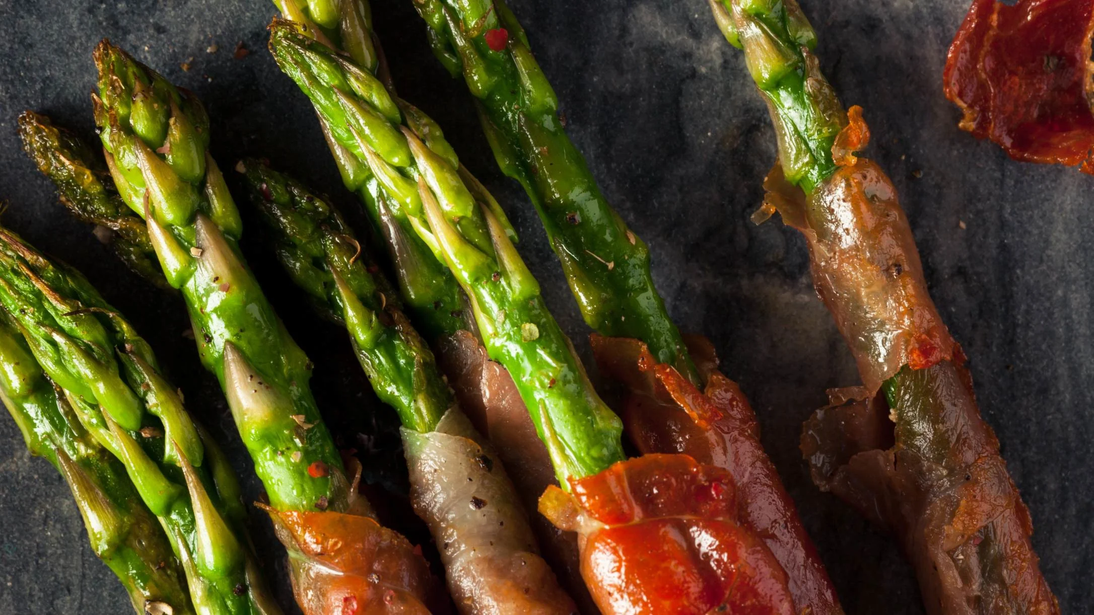 Roasted green asparagus spears wrapped in bacon with red pepper jelly glaze, arranged on a dark slate surface.