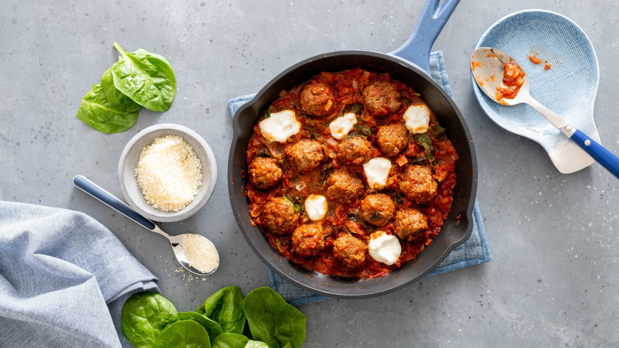 Cast iron skillet with meatballs in red tomato sauce topped with ricotta cheese, surrounded by spinach and parmesan.
