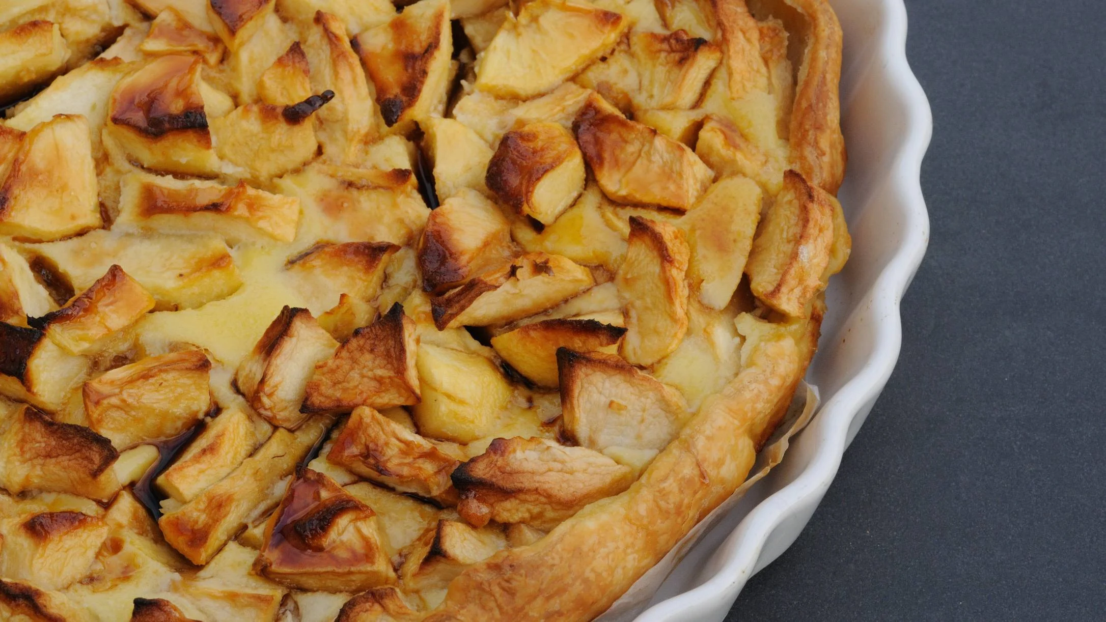 A golden-brown baked apple tart with caramelized apple chunks in a white ceramic scalloped pie dish on a gray surface.