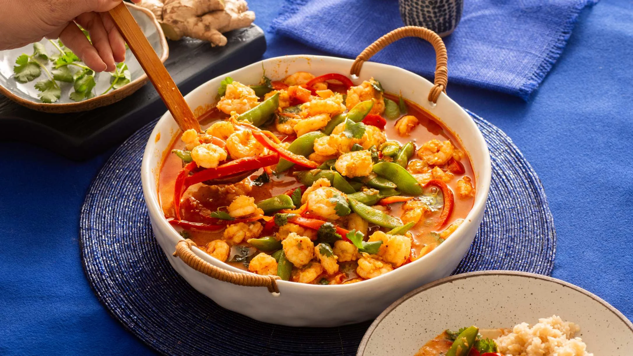 Spicy shrimp curry with red peppers and snap peas in a white serving bowl with wooden spoon on blue placemat.