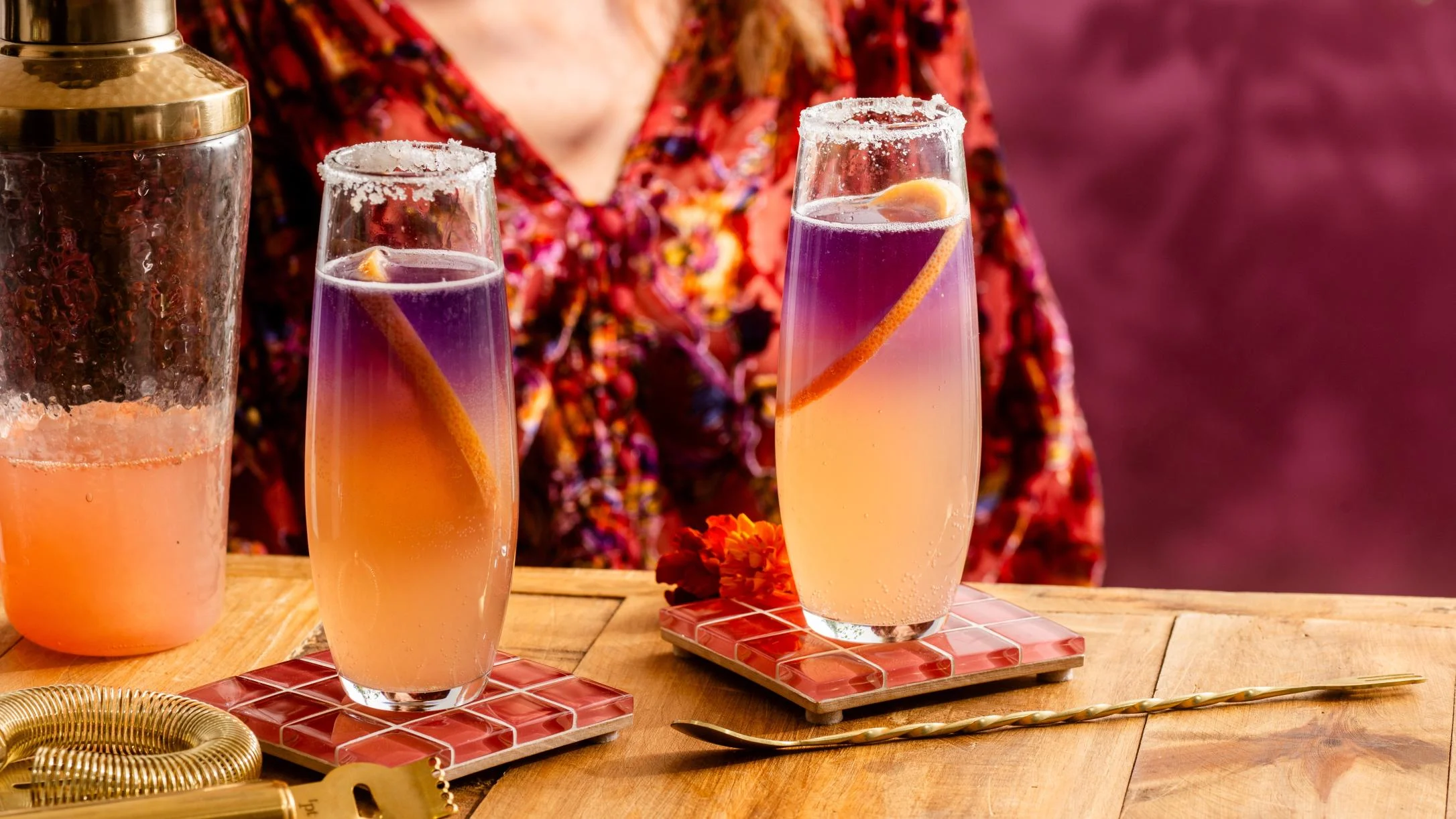 Two gradient purple-orange cocktails with sugar rims and citrus garnish on tile coasters beside a cocktail shaker.