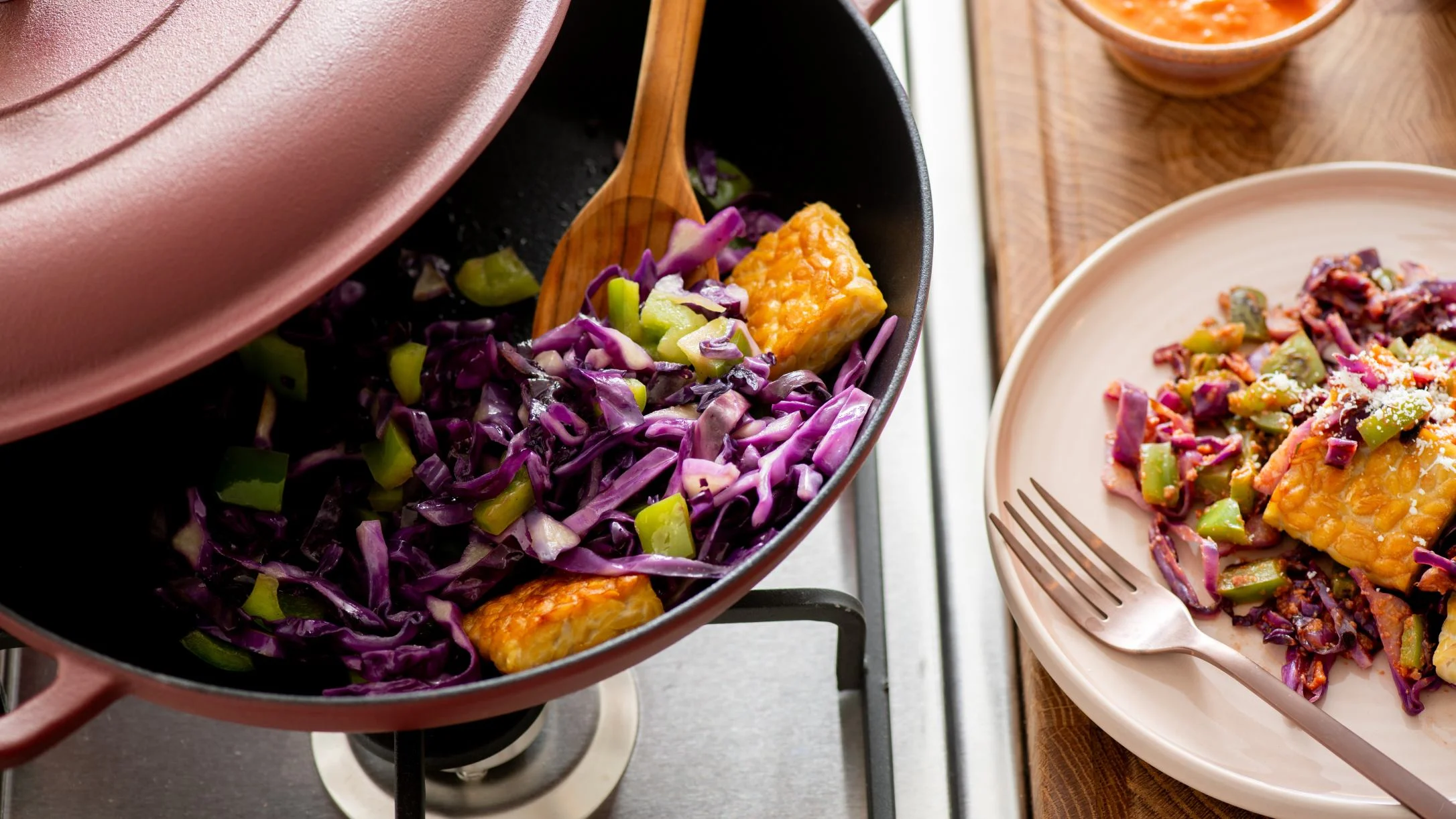 A black wok with purple cabbage, green peppers, and golden tempeh on a stove, beside a plated serving with a fork.