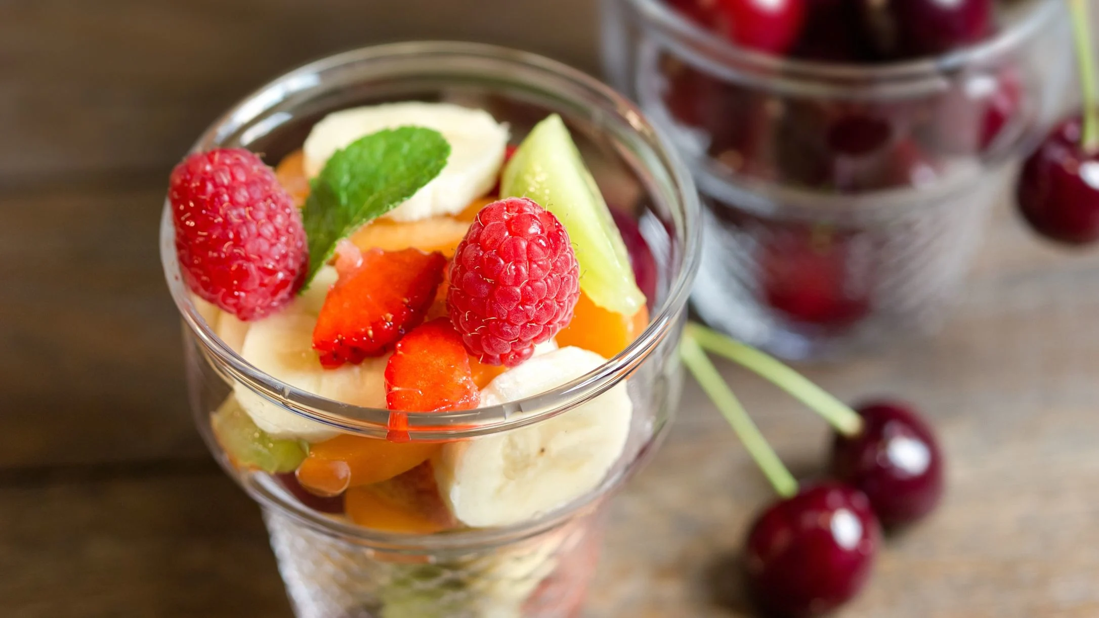 A glass cup filled with fresh fruit salad including raspberries, strawberries, banana, and kiwi, with cherries nearby.