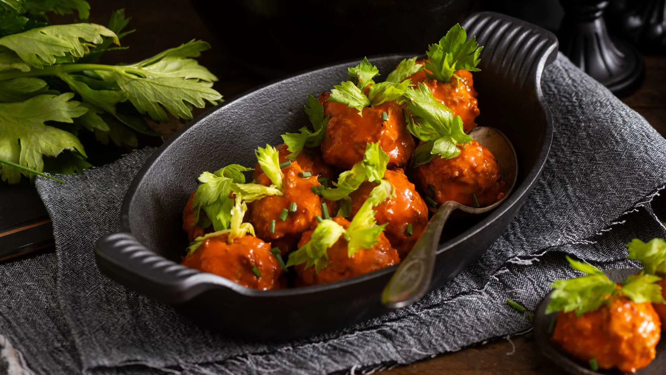 Meatballs in bright orange sauce garnished with fresh celery leaves, served in a cast iron dish on gray cloth.