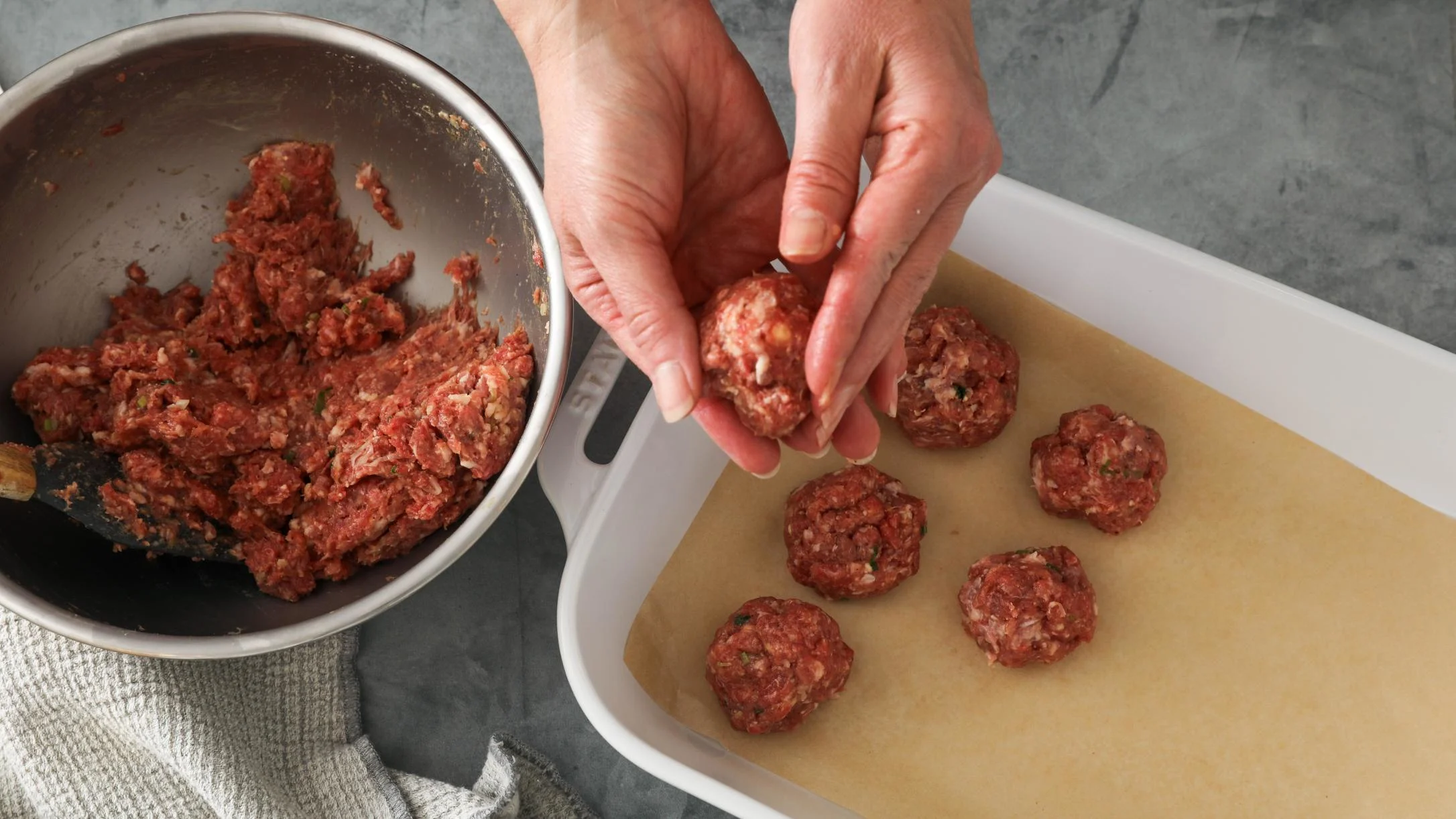Hands forming raw meatballs from ground beef mixture in metal bowl, placing them on parchment paper in white tray.