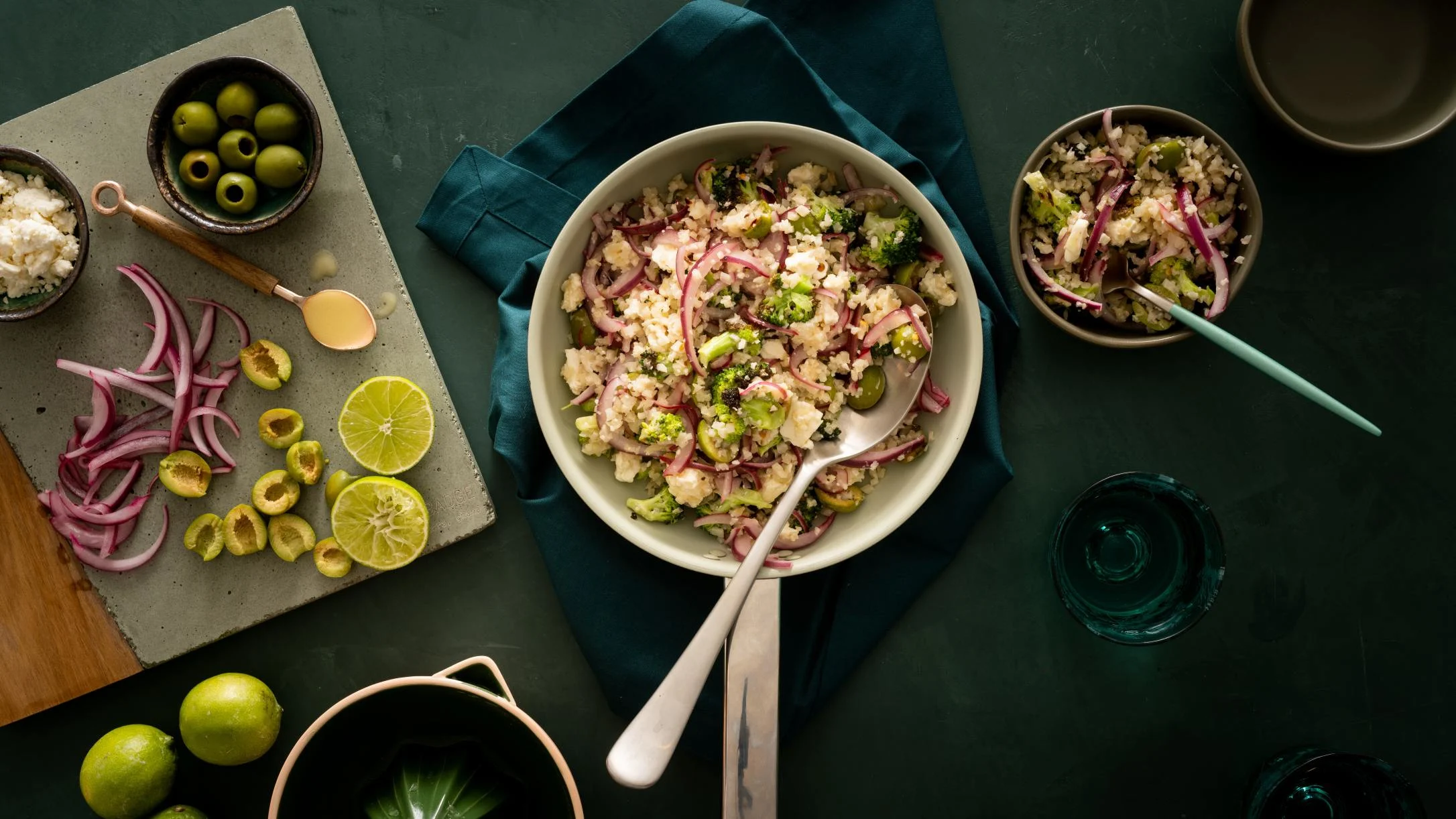 Skillet with cauliflower rice, broccoli and red onions, surrounded by green olives, lime slices, and ingredients on dark table.