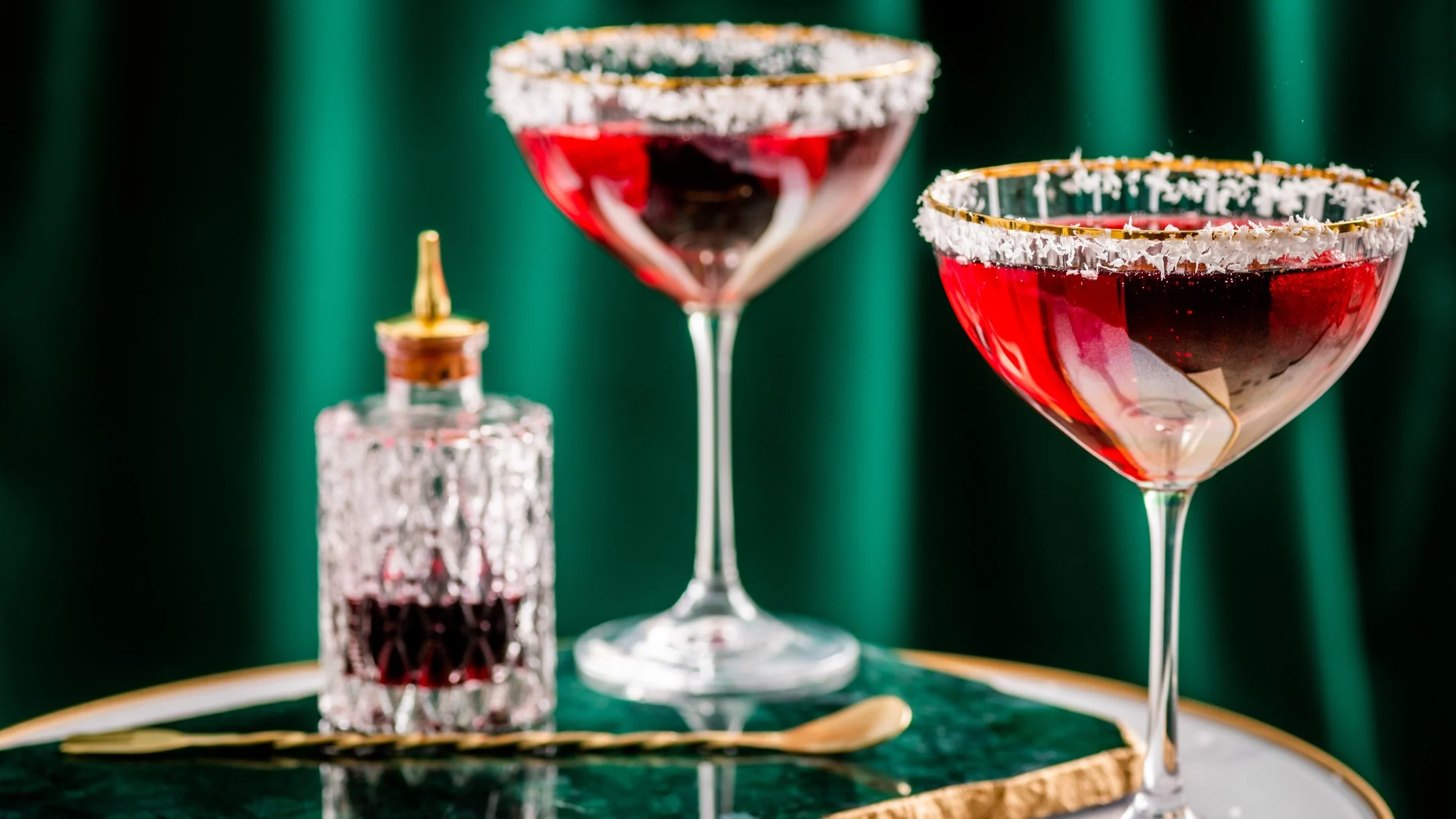 Two red cocktails with coconut-rimmed glasses on a green table, alongside a crystal bottle.