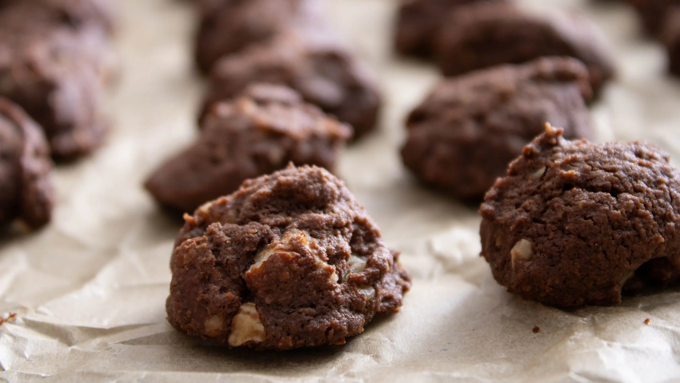 Freshly baked chocolate cookies with nut chunks arranged on parchment paper, with soft texture and rich brown color.