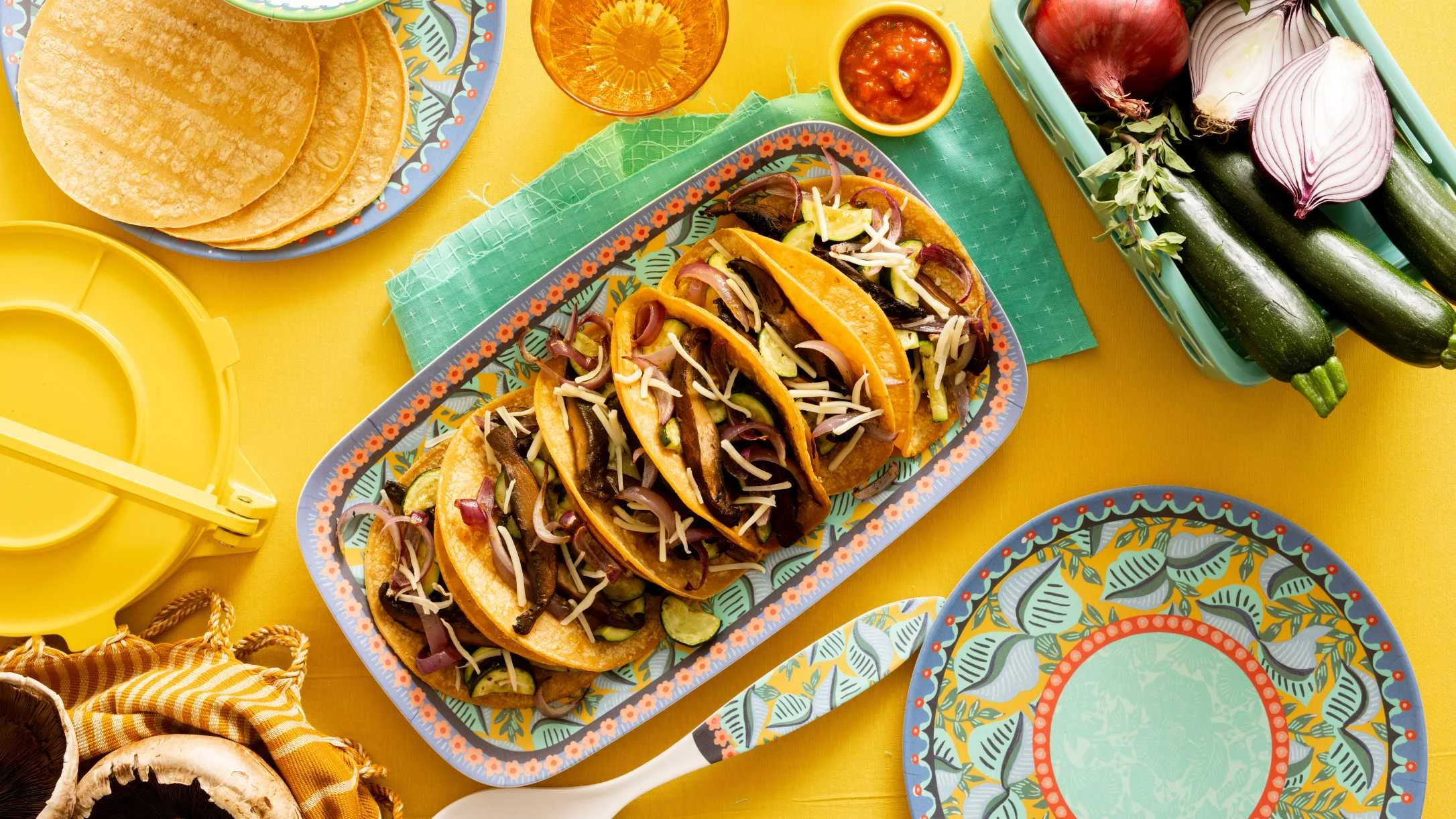 Colorful veggie tacos with zucchini, mushrooms, red onion, and shredded cheese on a decorative platter with salsa and tortillas.