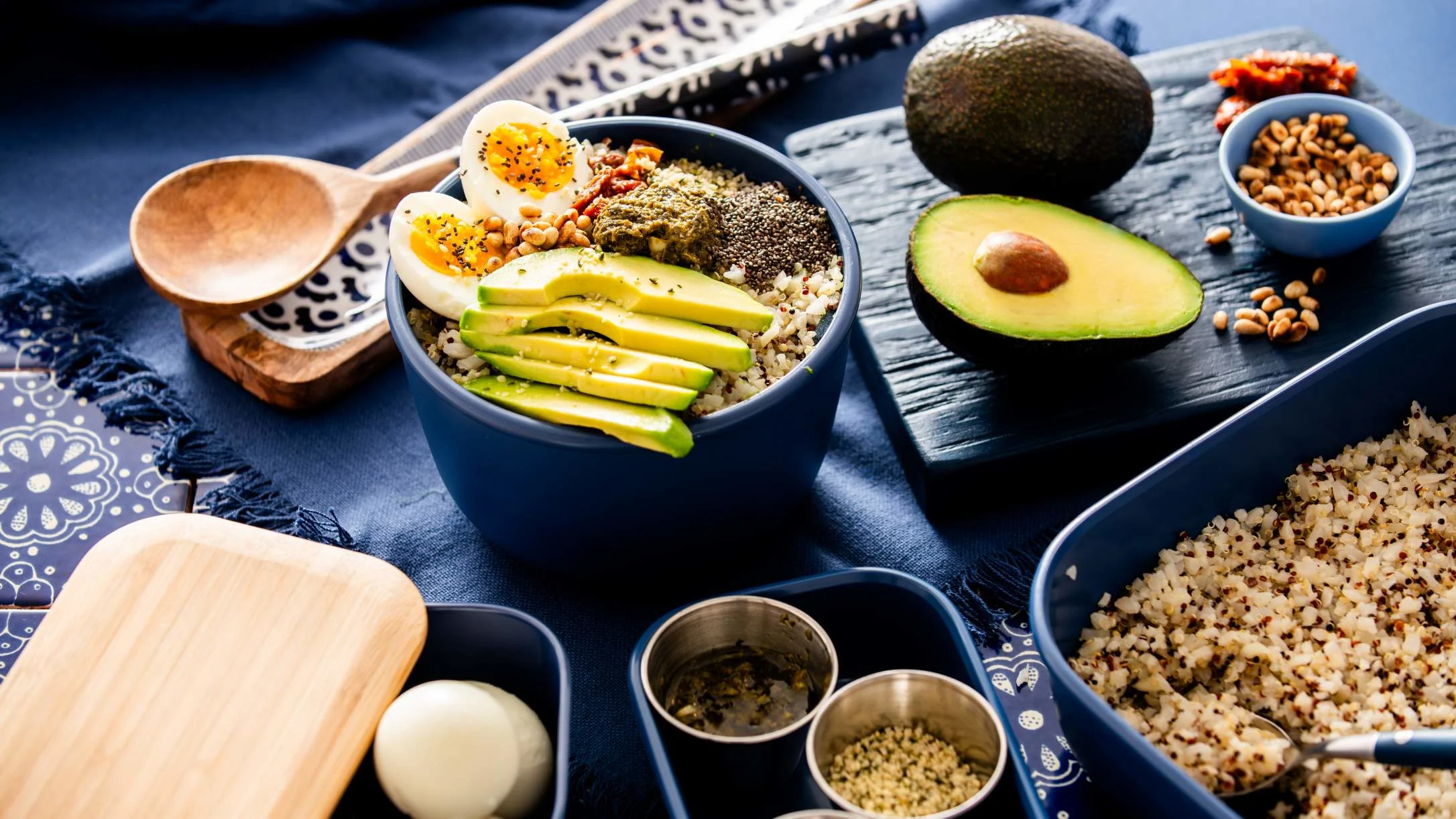 Healthy bowl with sliced avocado, boiled egg, quinoa, and pine nuts alongside wooden utensils on blue background.