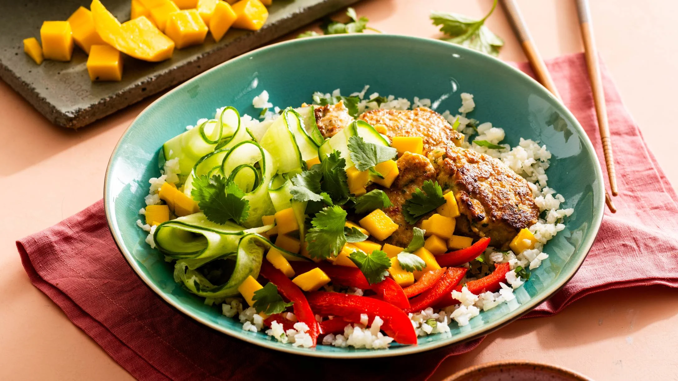 A teal bowl with cauliflower rice, grilled chicken, mango chunks, red pepper strips, cucumber ribbons, and cilantro on a red napkin.