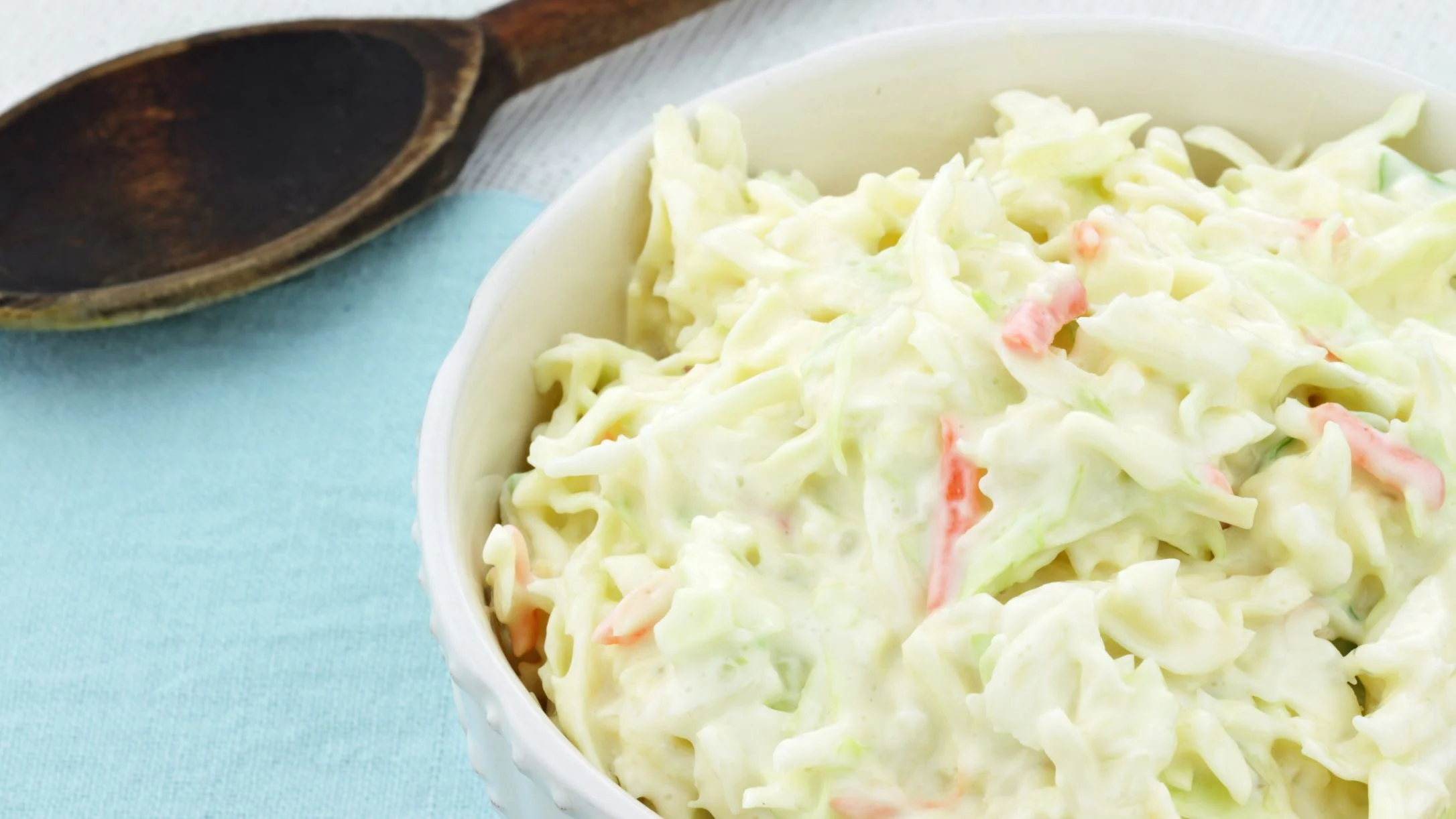 Creamy coleslaw in a white bowl with shredded cabbage, wooden spoon visible on light blue tablecloth.