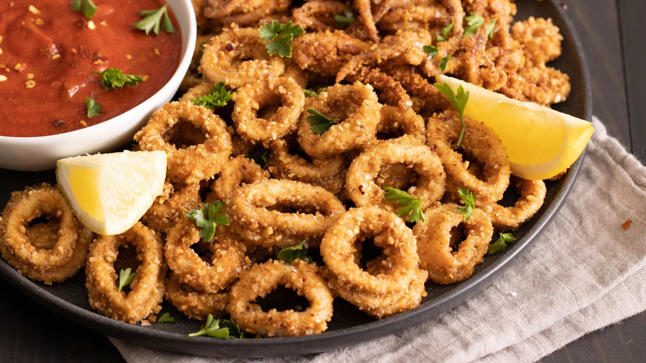 Crispy fried calamari rings on a dark plate with lemon wedges and marinara dipping sauce, garnished with parsley.