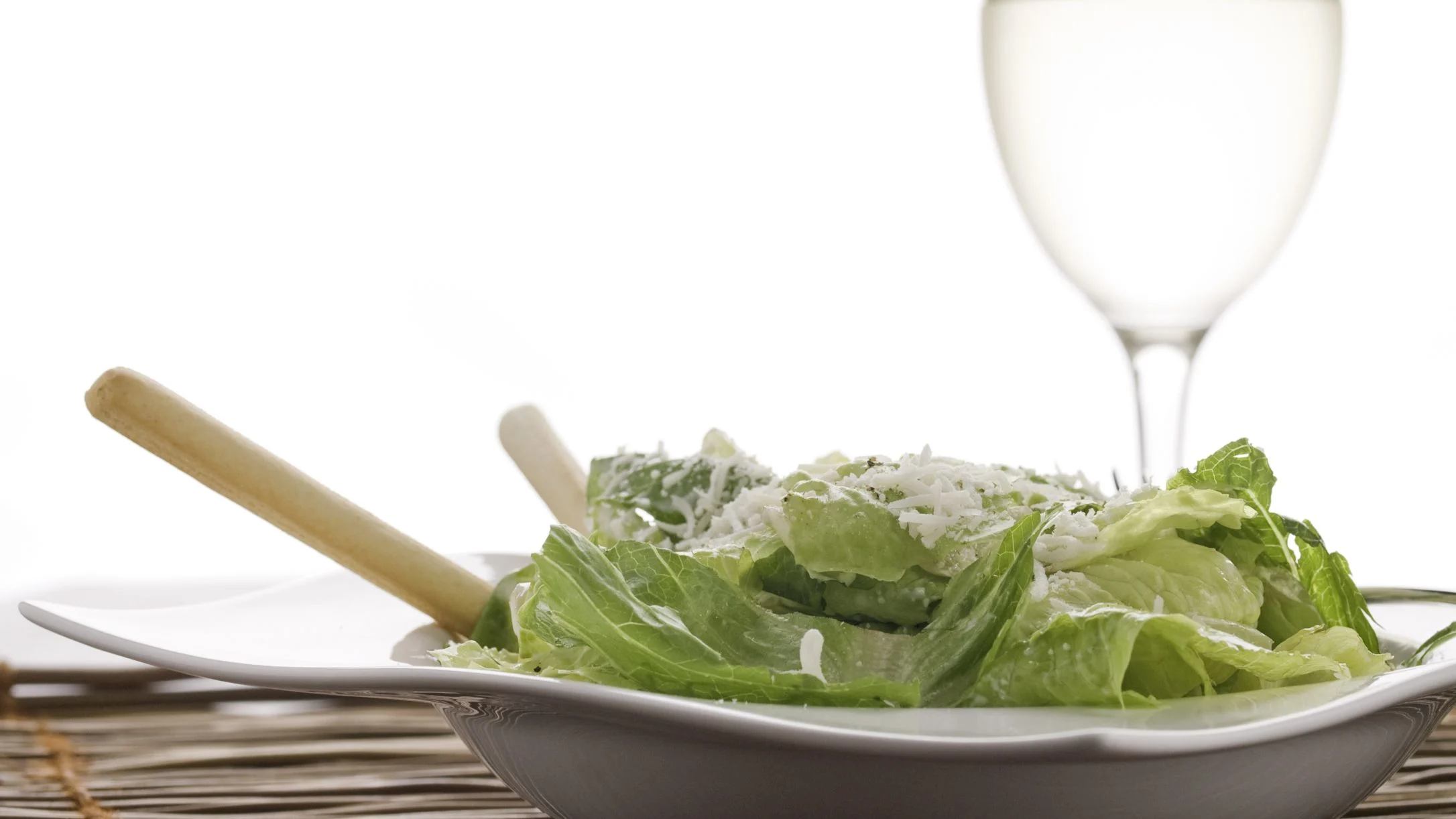 A white bowl of green Caesar salad topped with shredded Parmesan, with wooden serving utensils and a wine glass in the background.