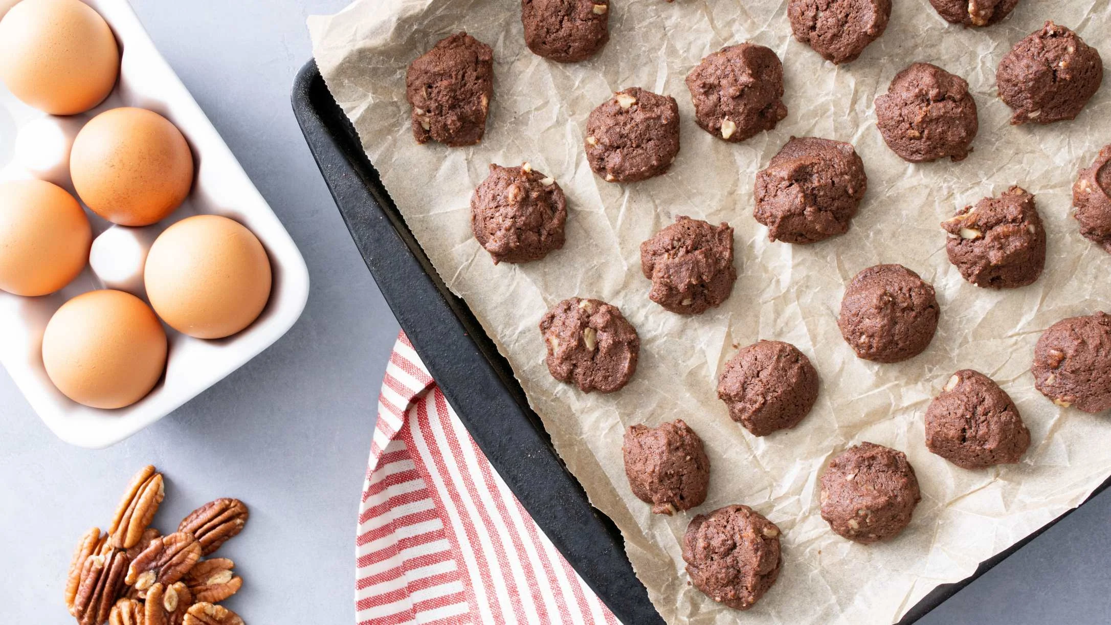Chocolate cookies on parchment paper with brown eggs in a dish and pecans on the side.