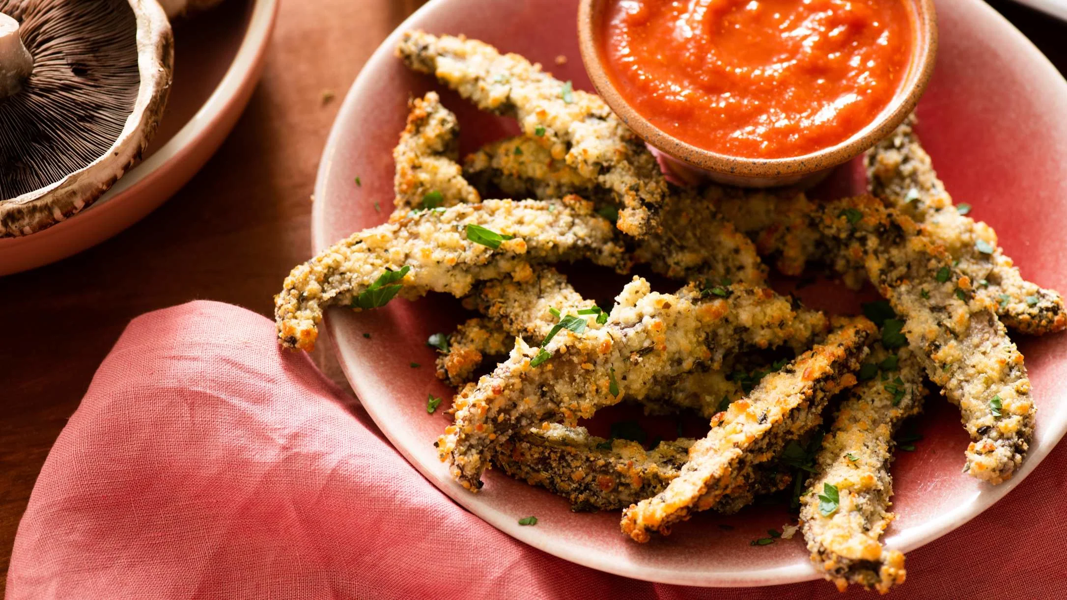 Crispy breaded mushroom fries on a pink plate with marinara dipping sauce, garnished with fresh herbs.