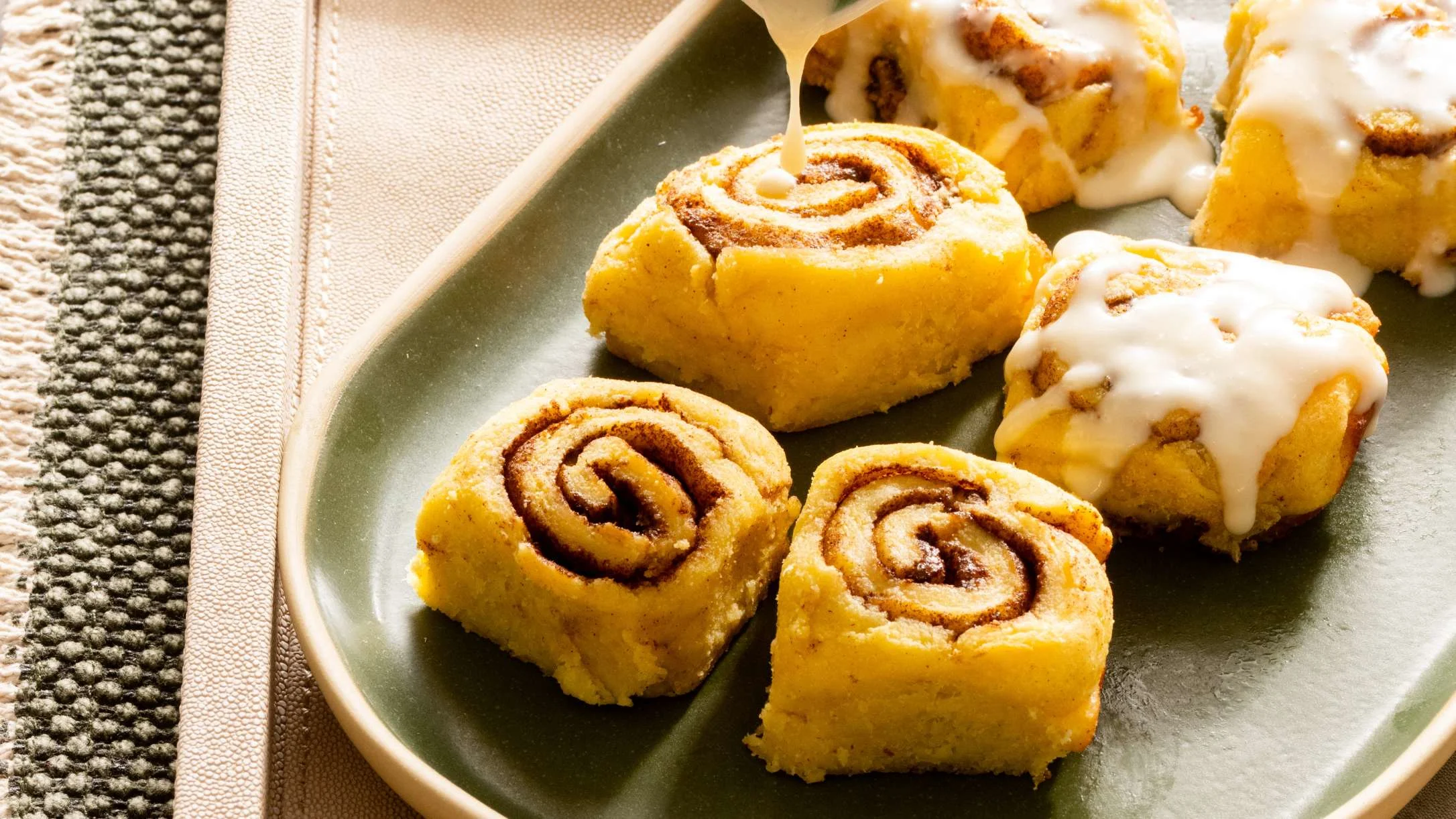 Golden cinnamon rolls on a green plate, with white cream cheese icing being drizzled over them from a spoon.