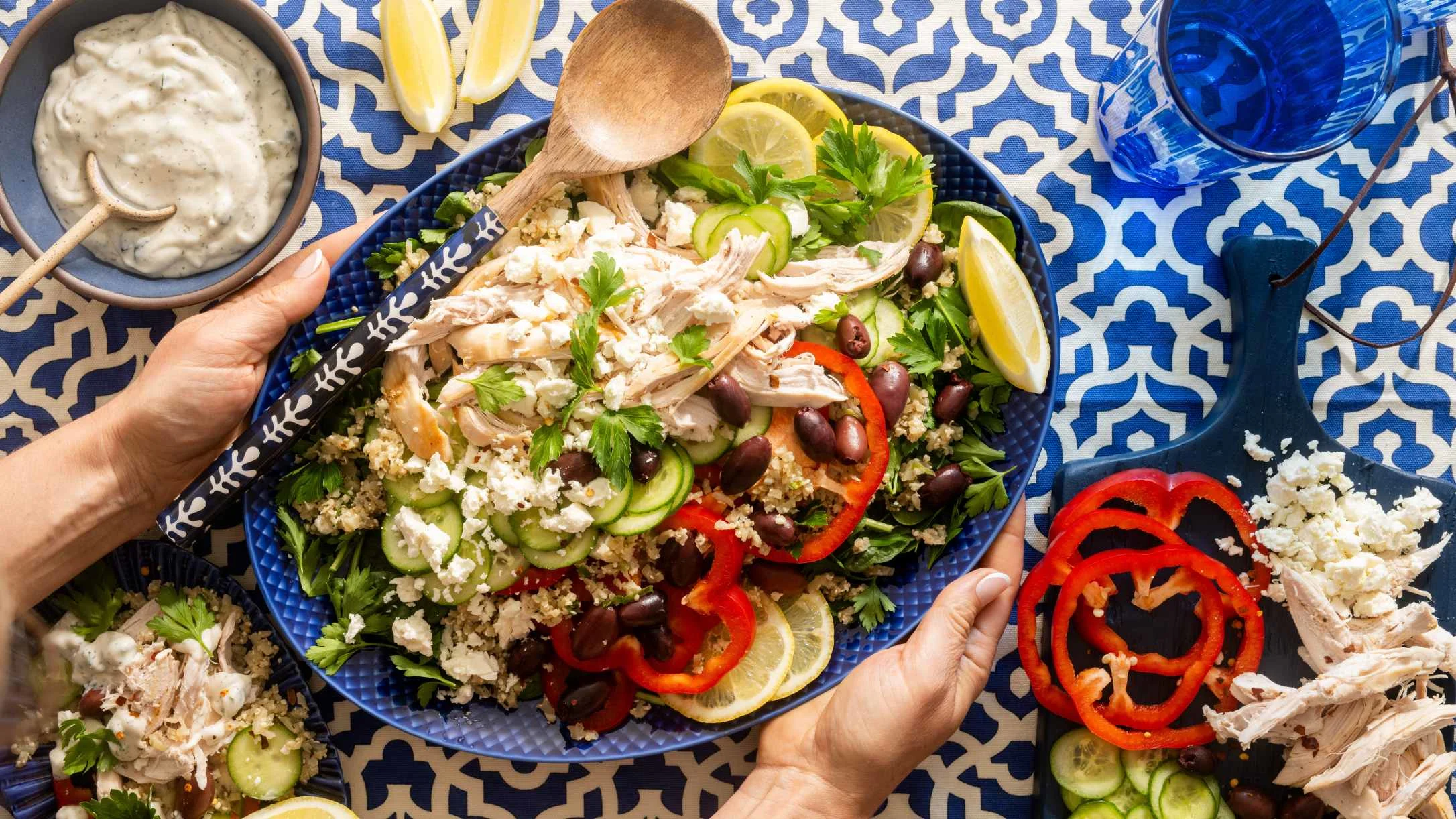 Hands holding a blue platter of Mediterranean salad with shredded chicken, feta, olives, red peppers, cucumber, and lemon.