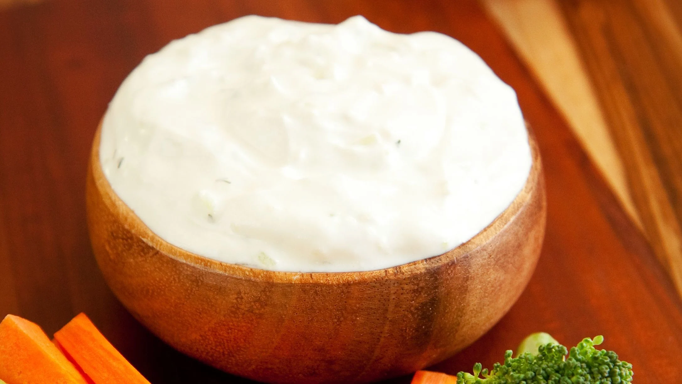 A wooden bowl filled with creamy white dip, surrounded by fresh carrot sticks and broccoli on a wooden surface.