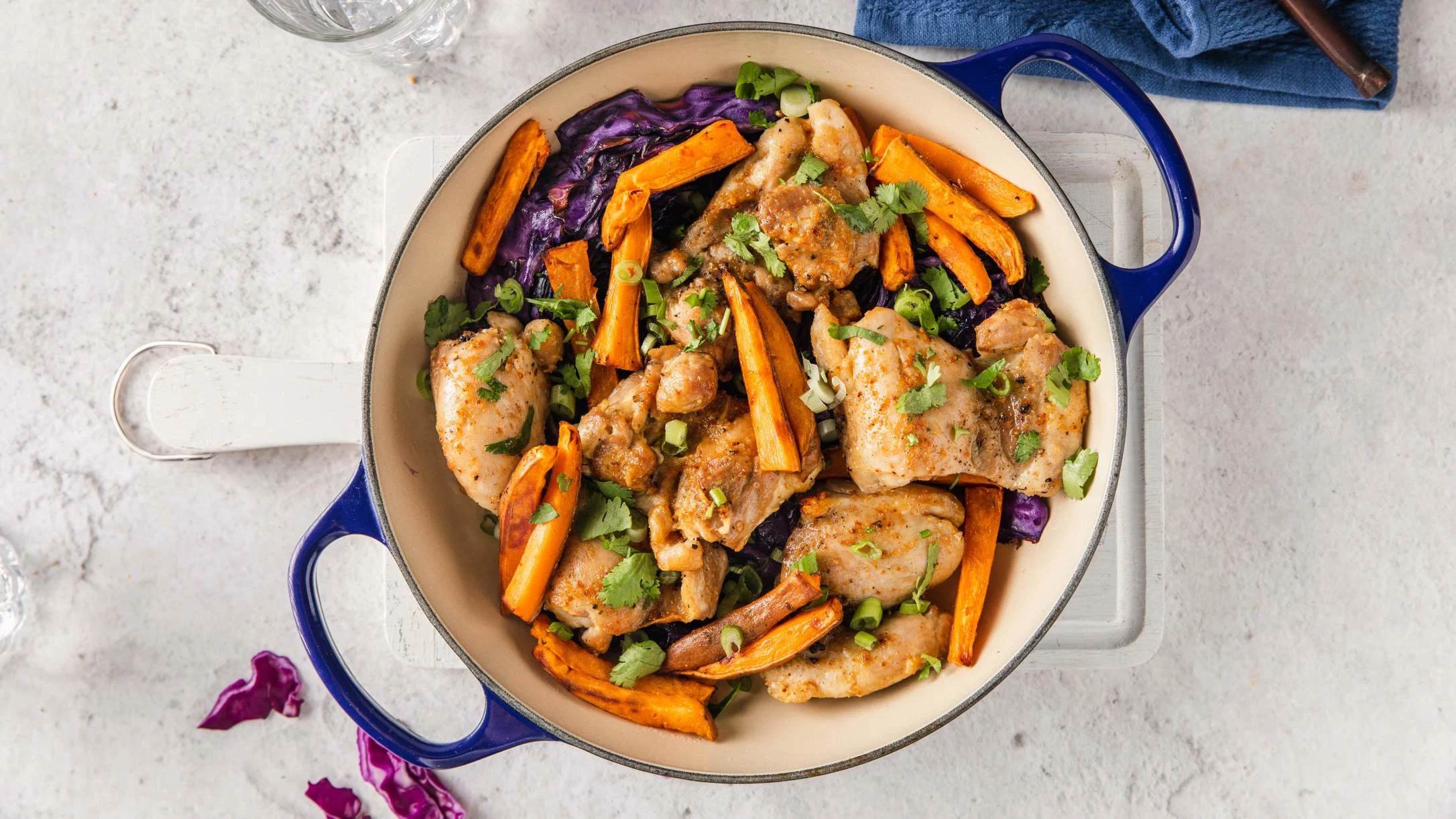 Blue cooking pot containing roasted chicken pieces with sweet potato, purple cabbage, and fresh herbs on a white surface.