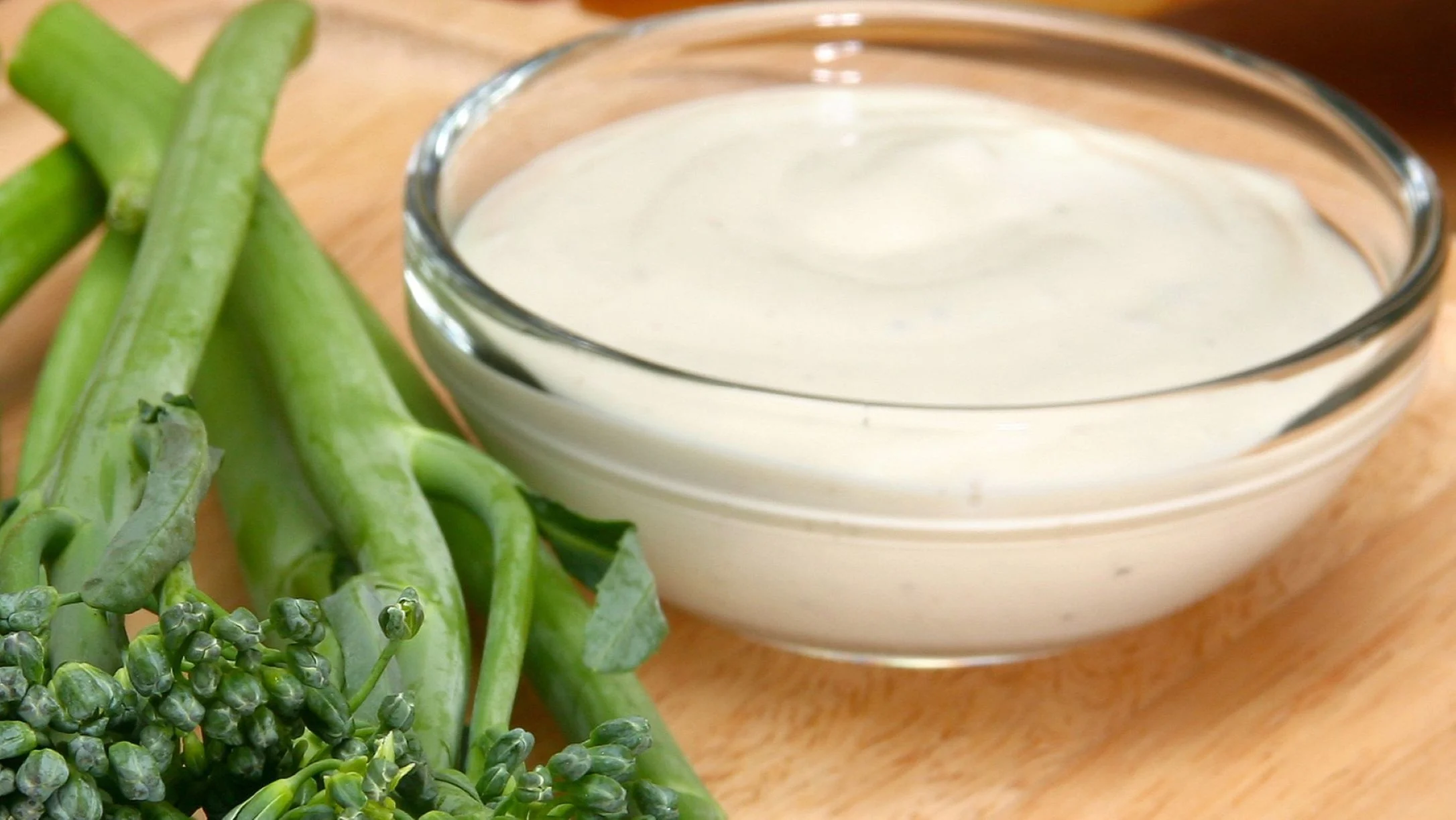 Fresh green broccoli next to a glass bowl of creamy white ranch dressing on a wooden cutting board.