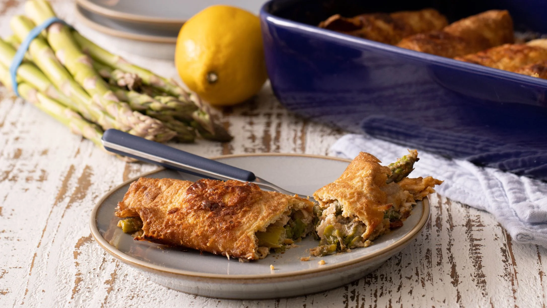 A golden-baked asparagus crepe cut open on a gray plate, with fresh asparagus, a lemon, and a blue baking dish nearby.