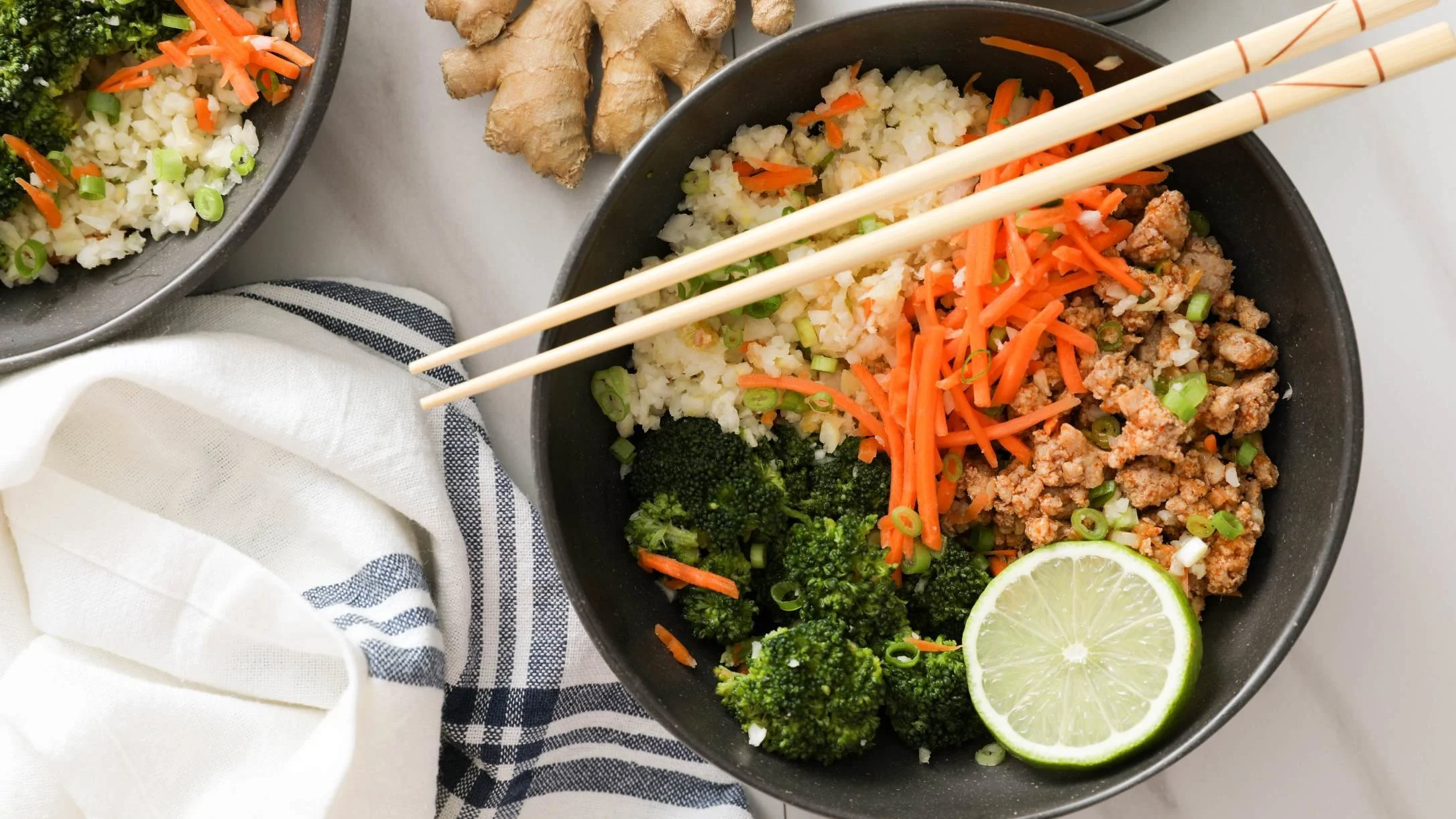 A dark bowl with cauliflower rice, ground meat, shredded carrots, broccoli, and lime, topped with chopsticks, beside fresh ginger.