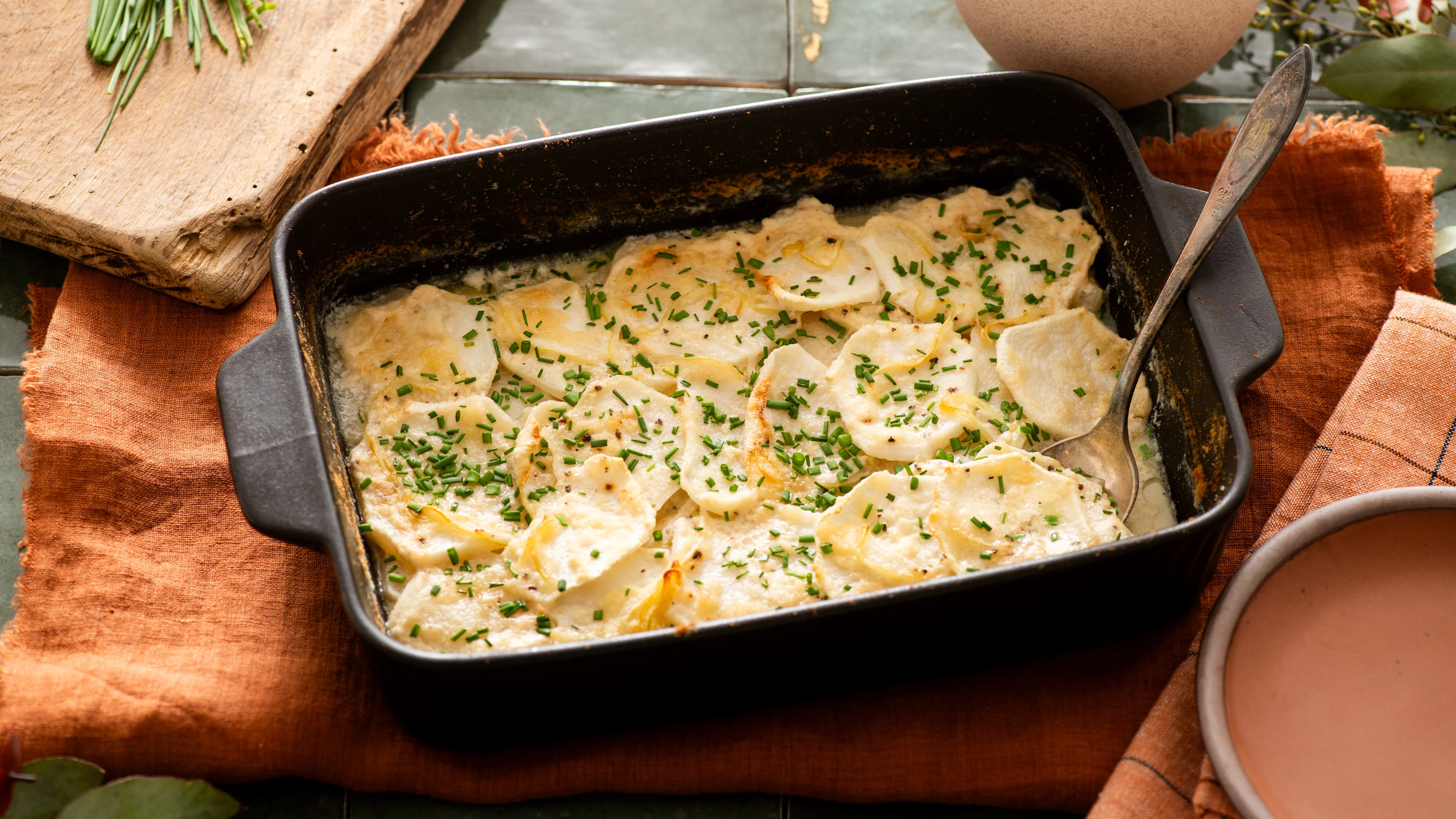Creamy faux potato gratin in black baking dish with fresh chives, served on orange linen with serving spoon.