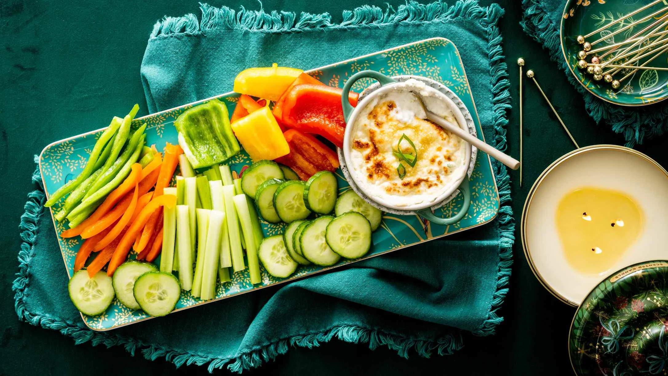 Colorful vegetable platter with cucumber, carrots, and bell peppers served with baked dip on a teal decorative tray.