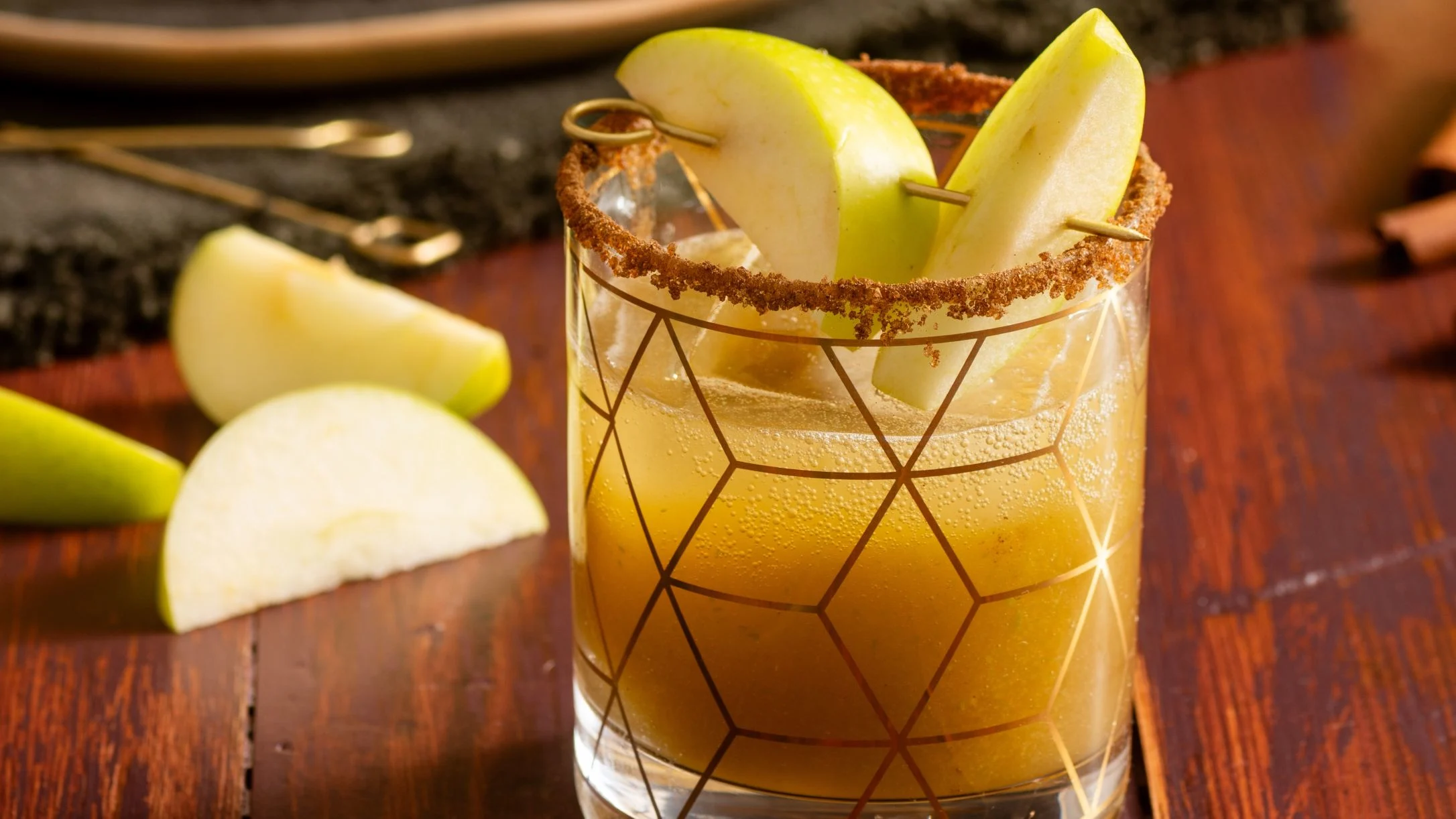 A golden apple cocktail in a geometric glass with a spiced rim, garnished with fresh green apple slices on a wooden surface.
