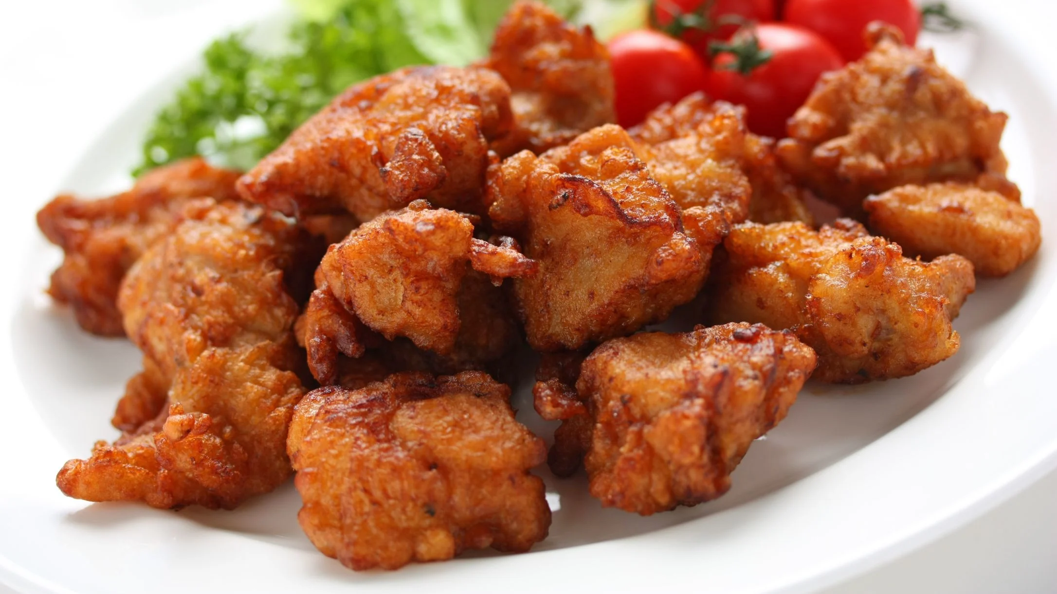 Golden-brown crispy fried chicken pieces on a white plate, garnished with fresh cherry tomatoes and green lettuce leaves.
