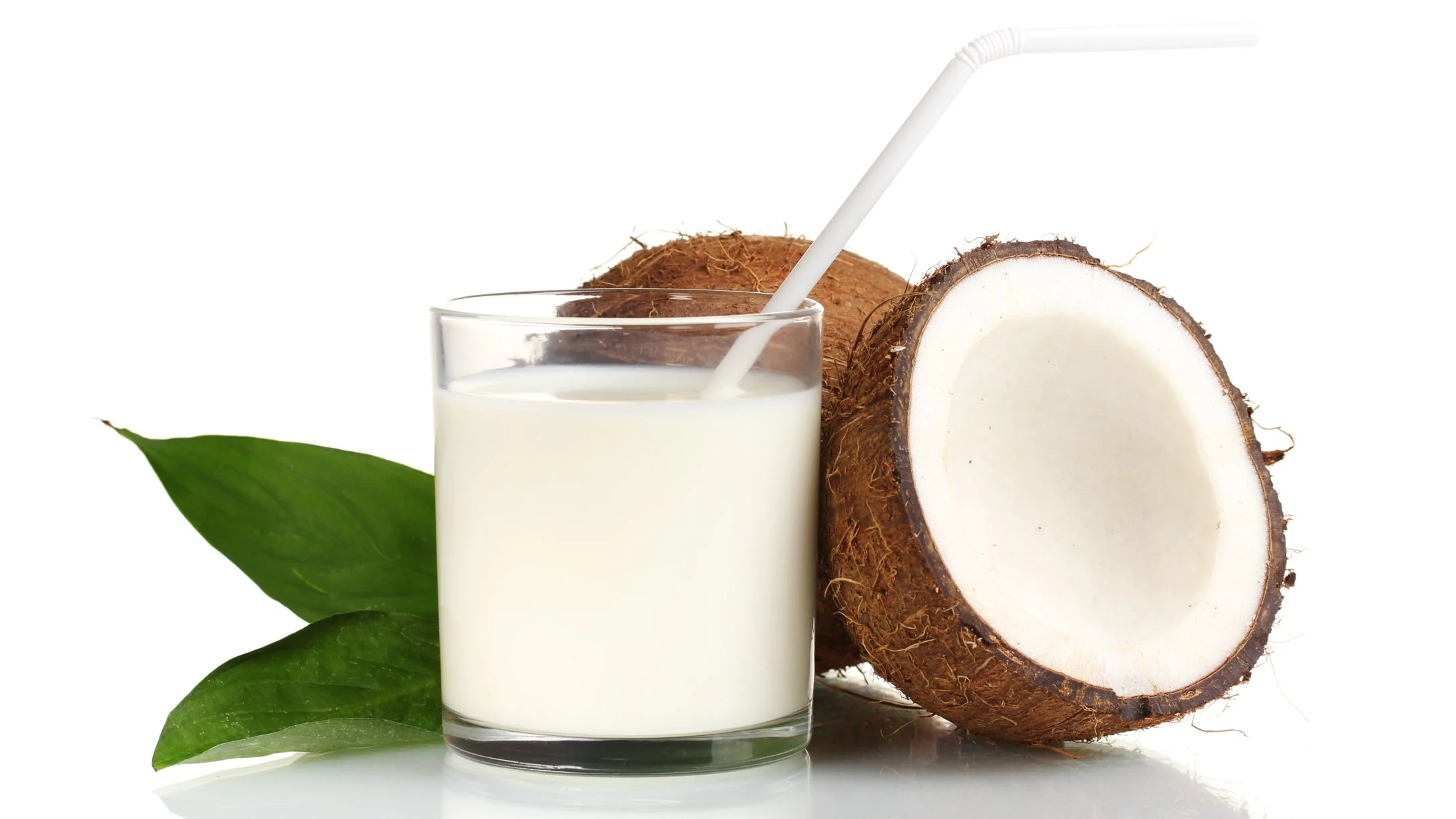 A glass of coconut shake with a straw beside a halved coconut and green leaves on a white background.