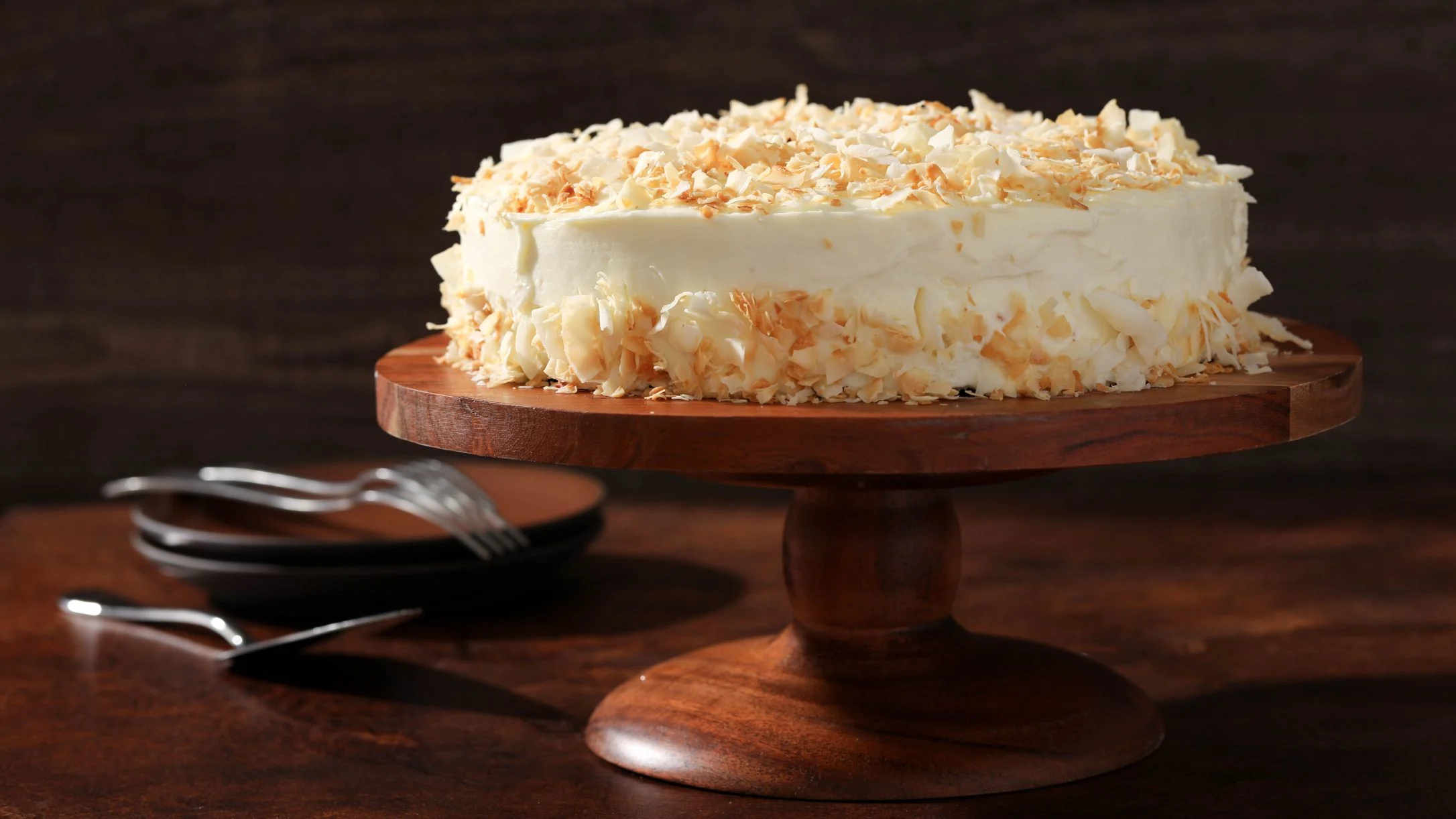 A coconut layer cake with white frosting and toasted coconut flakes on a wooden cake stand, with forks and plates nearby.