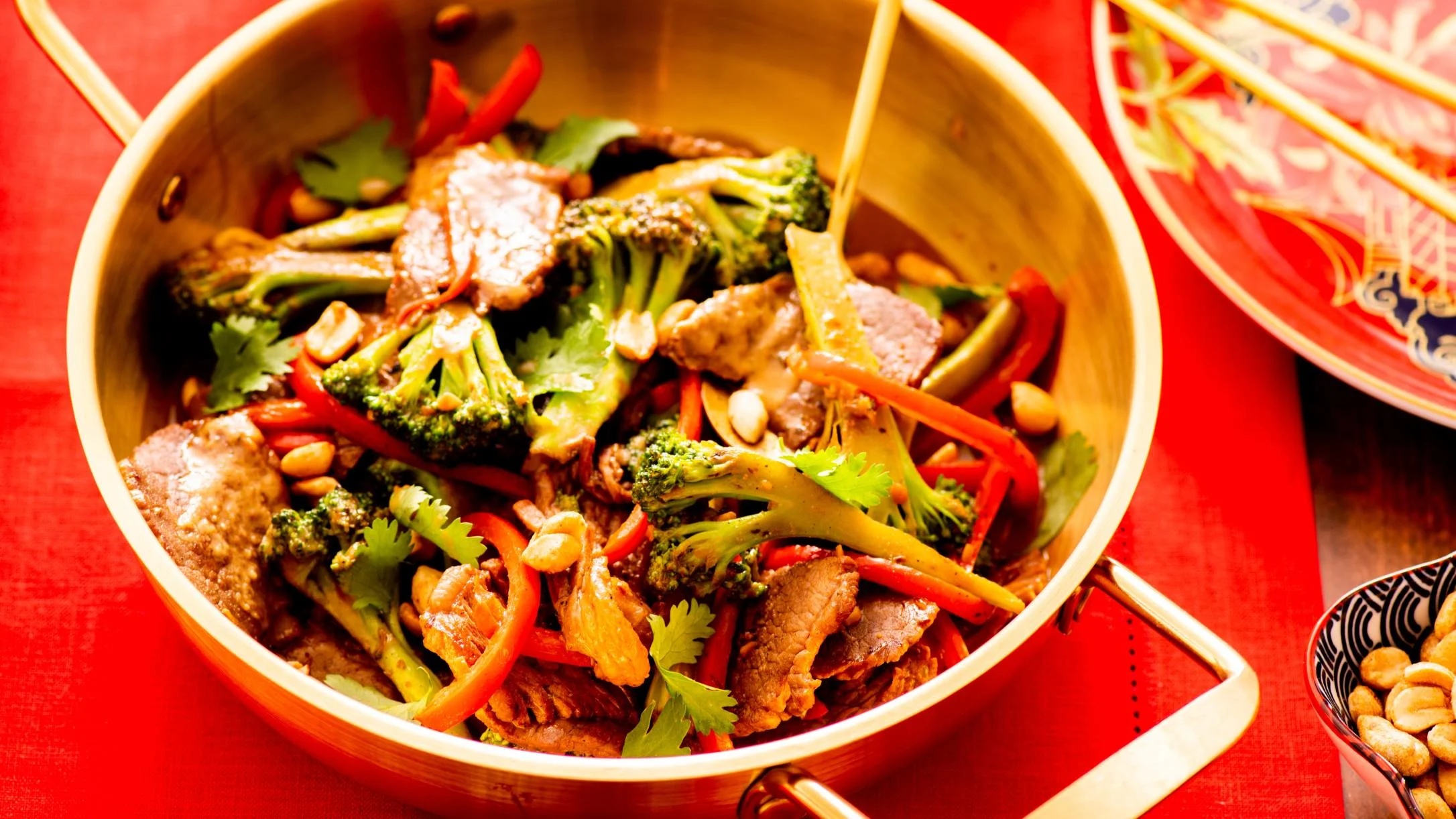 A gold wok filled with beef stir-fry featuring broccoli, red peppers, peanuts, and cilantro on a red surface.