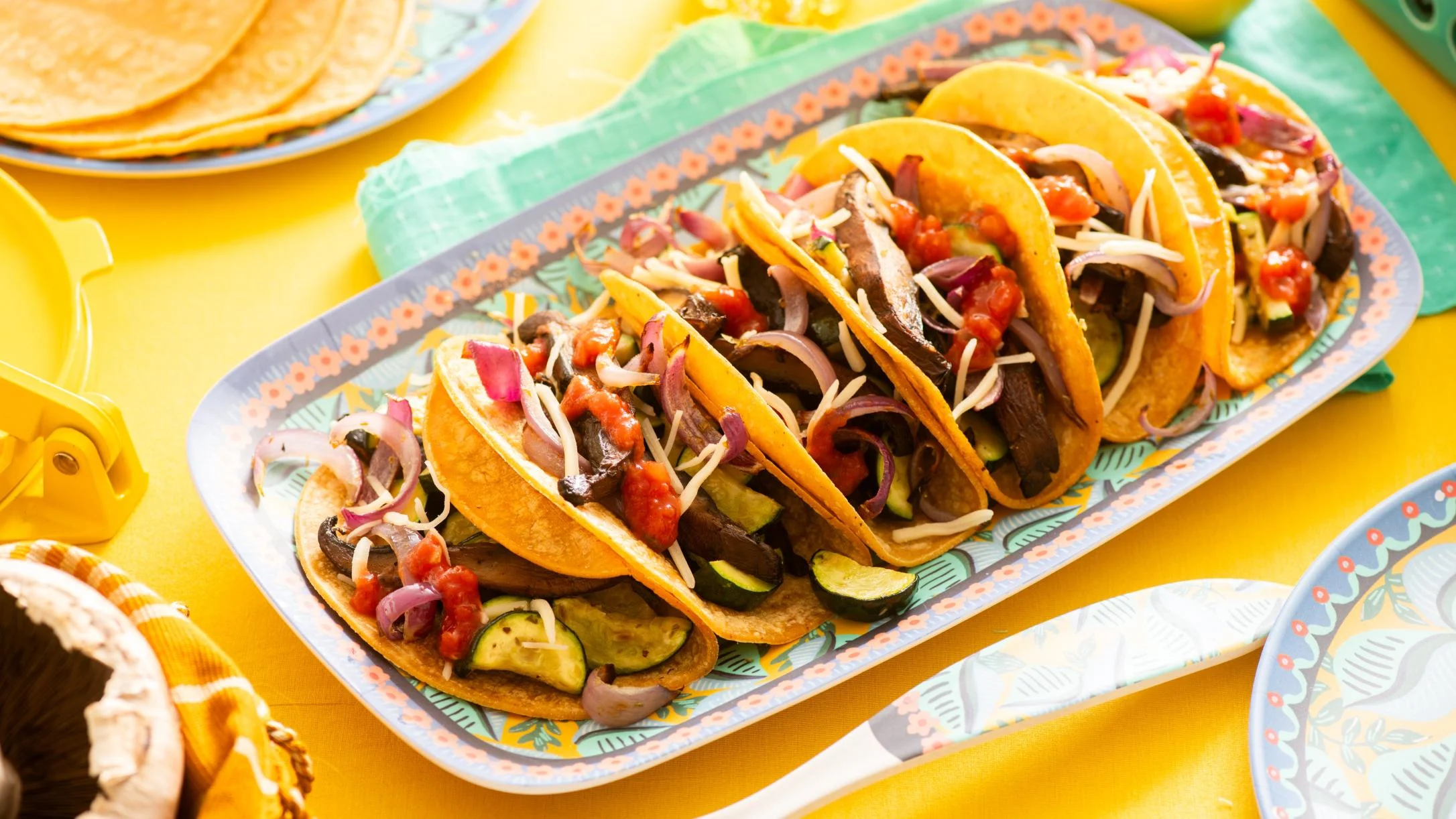 Four veggie tacos in yellow corn tortillas topped with zucchini, mushrooms, red onion, salsa, and shredded cheese on a colorful platter.