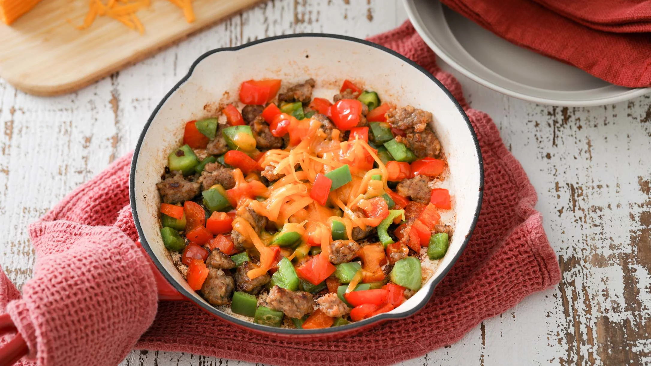 Skillet meal with sausage, diced red and green peppers, topped with melted orange cheese on a red towel.