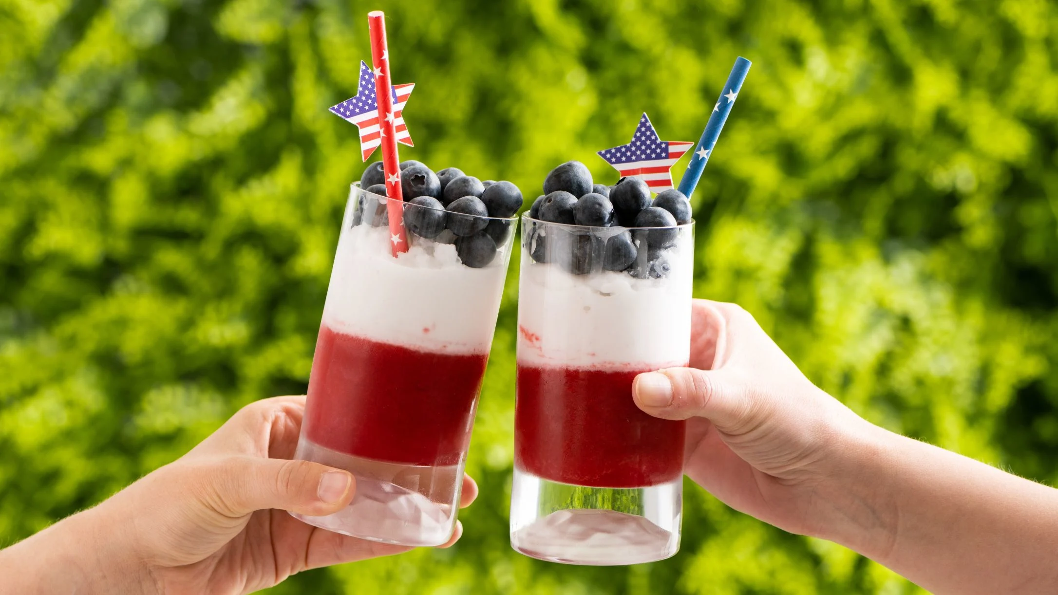 Two hands toasting patriotic red, white, and blue layered drinks with blueberries and American flag star decorations.