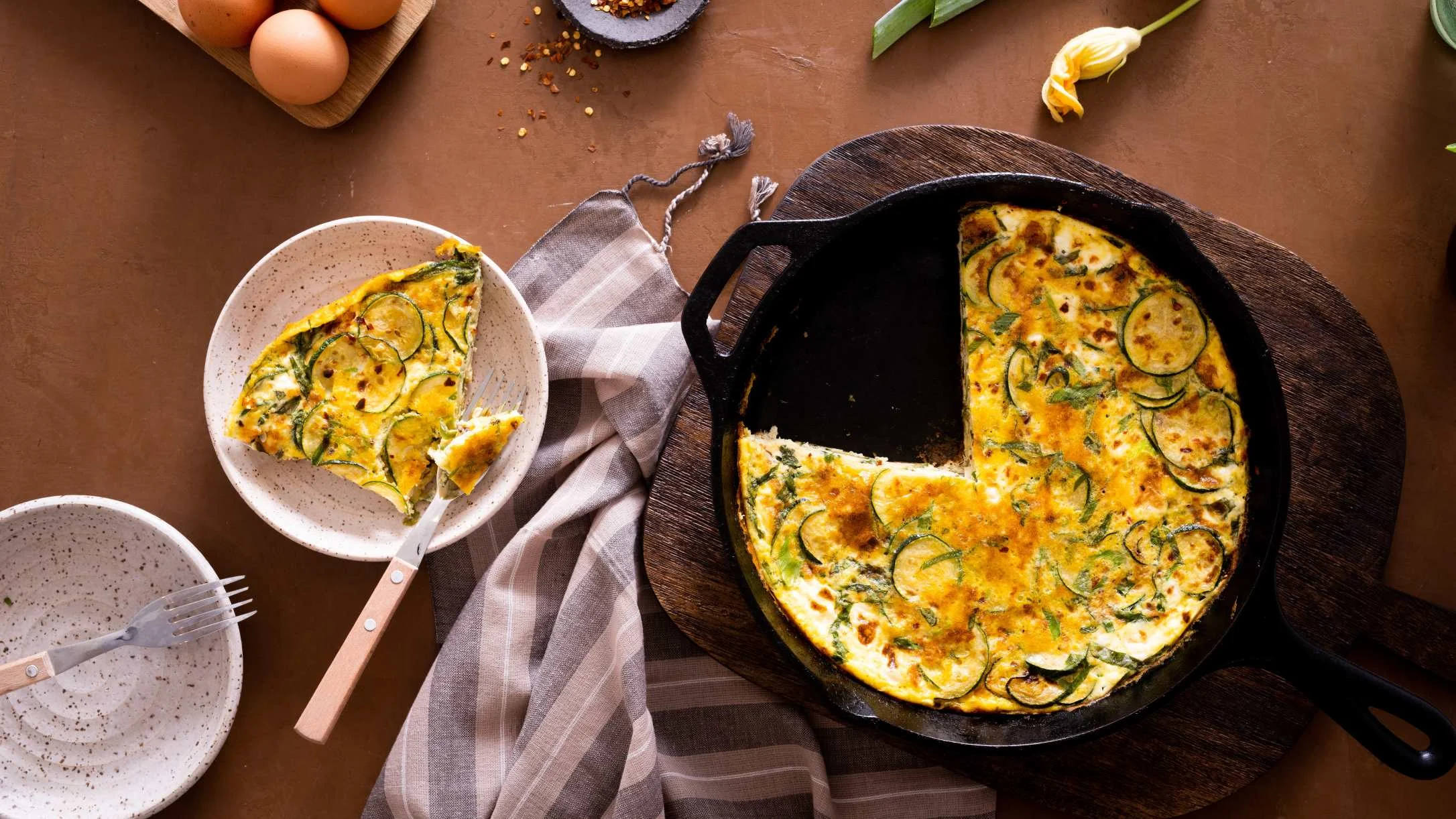 Zucchini frittata in cast iron skillet with slice served on plate, fresh eggs and spices visible on rustic brown surface.
