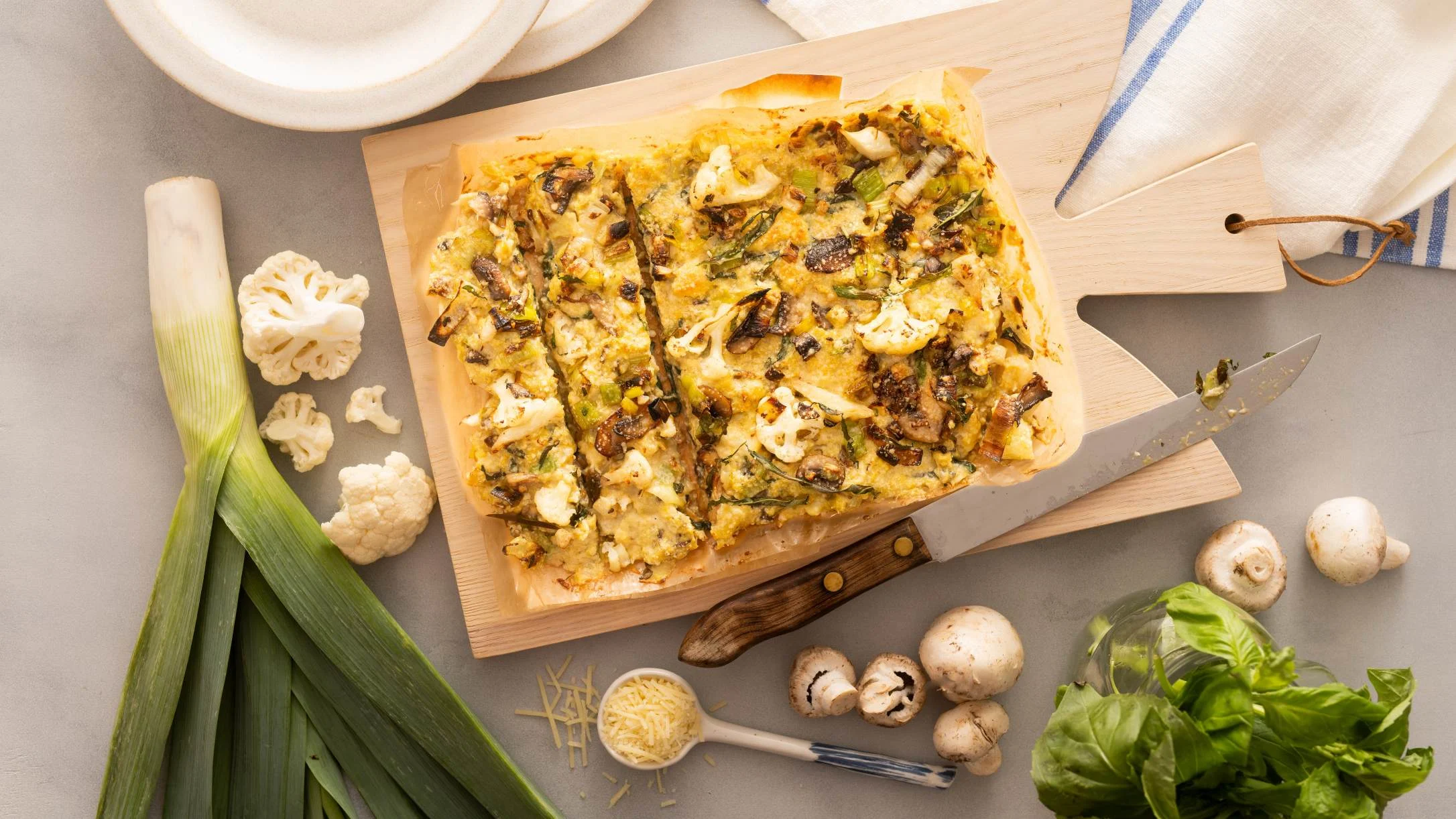 Sliced vegetable frittata on a wooden board, surrounded by leeks, cauliflower, mushrooms, basil, and shredded cheese.