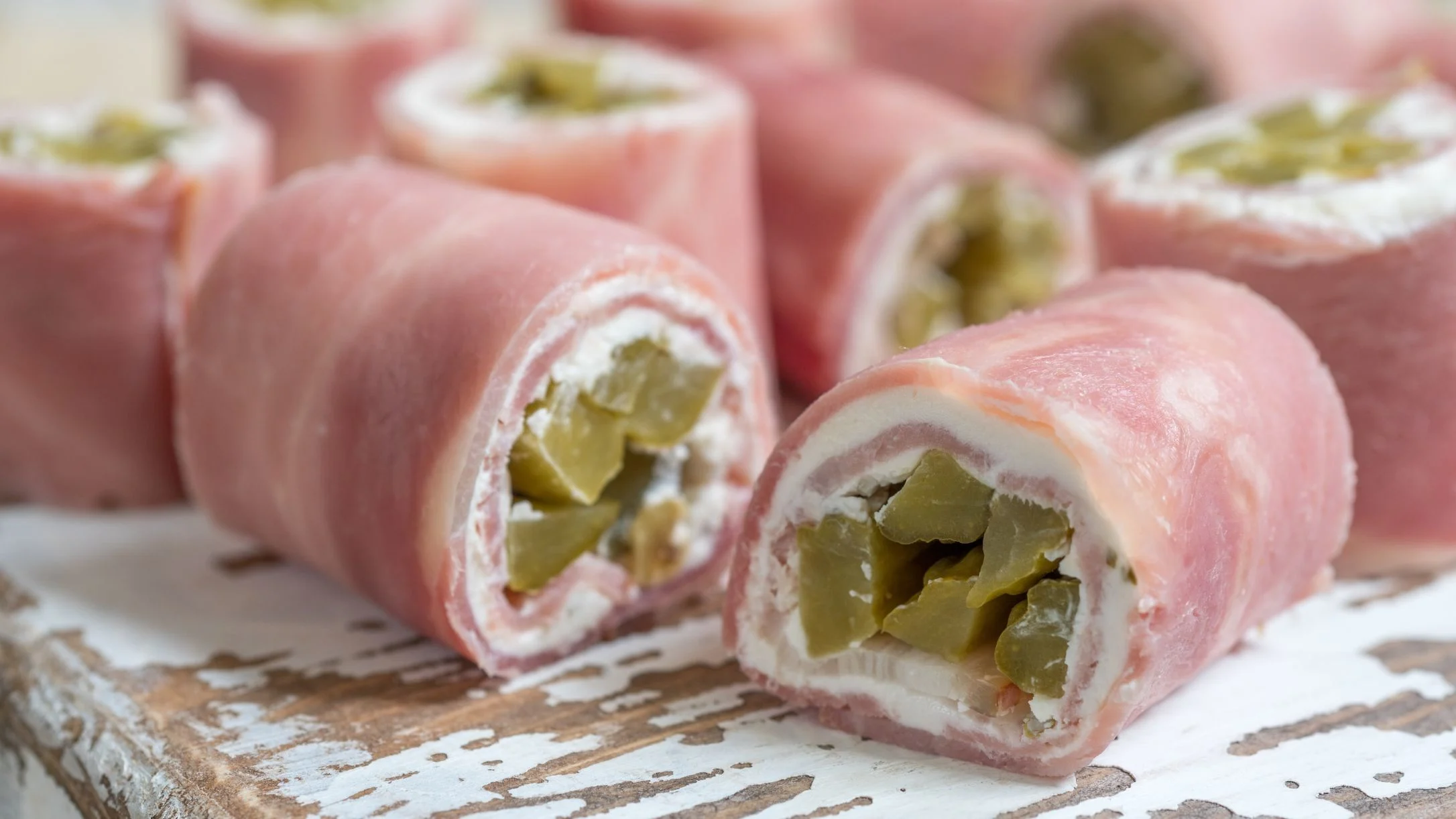 Vegan ham roll-ups filled with cream cheese and diced pickles, arranged on a rustic white wooden board.