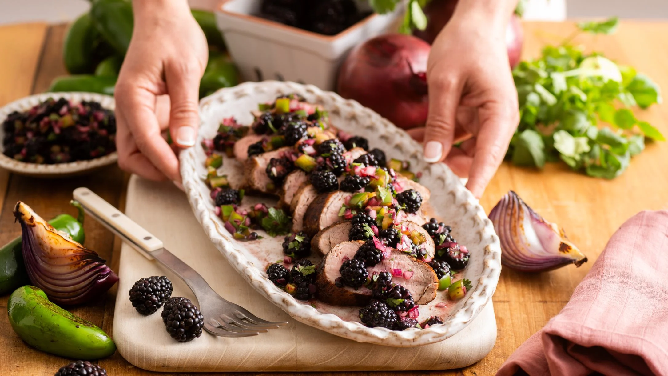 Hands holding a white serving dish of sliced pork tenderloin topped with blackberry salsa, surrounded by fresh ingredients.