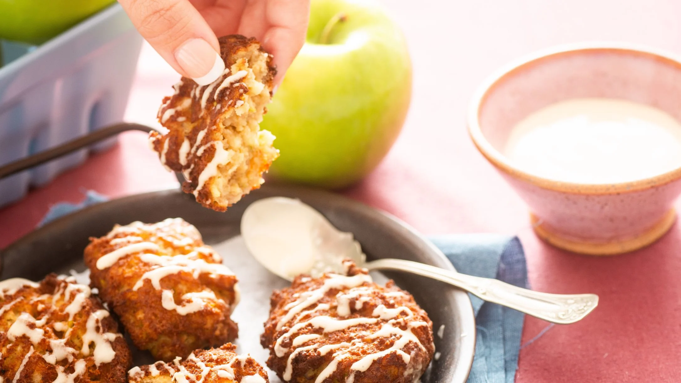 A hand picks up a glazed apple fritter from a skillet. A green apple and bowl of glaze sit nearby.