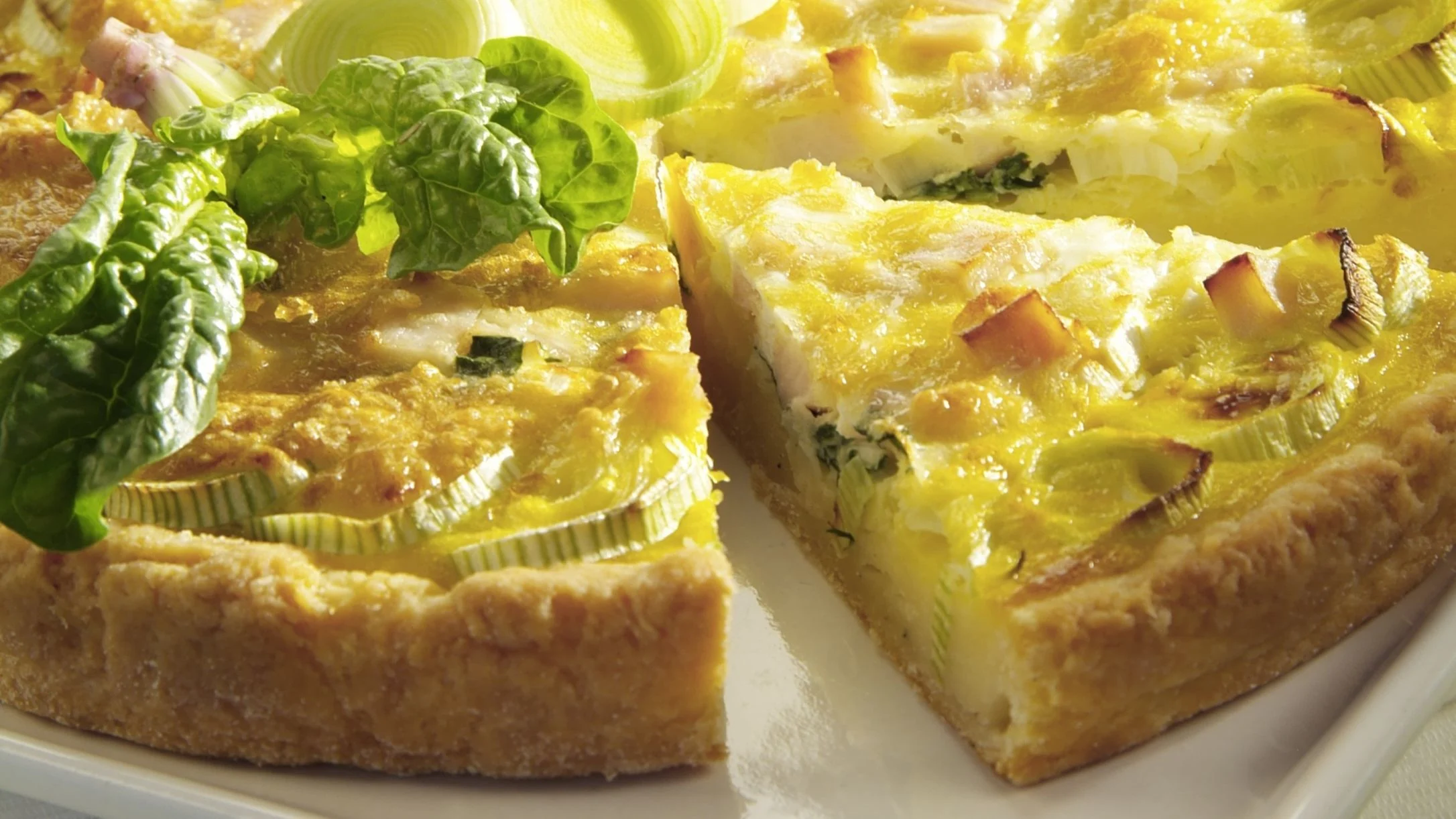 A golden leek and cheese quiche with a flaky crust, one slice pulled apart, garnished with fresh spinach leaves.