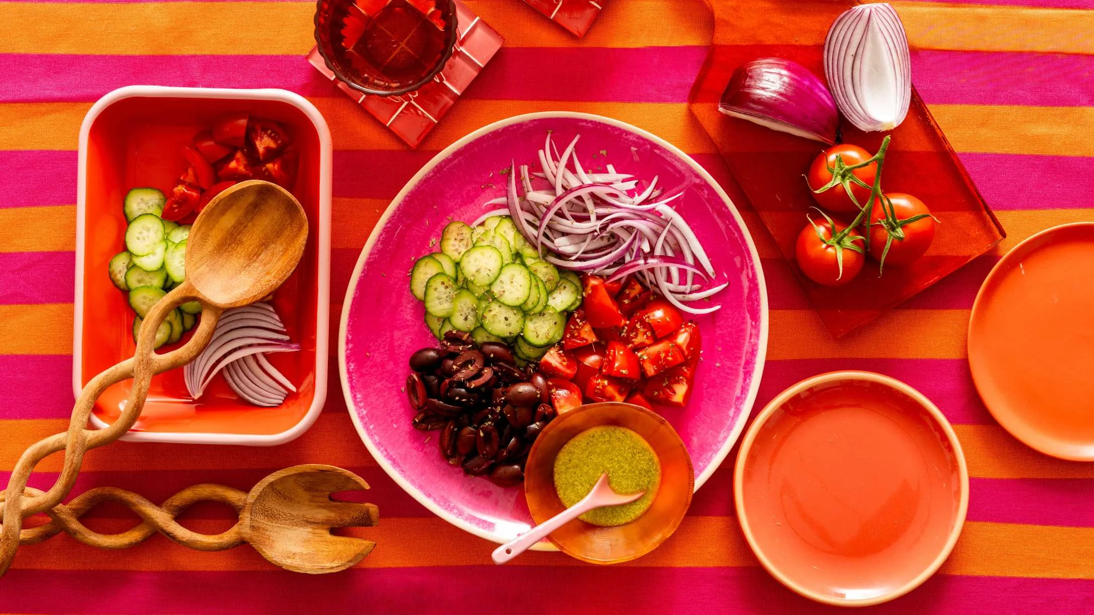 A vibrant salad with cucumbers, tomatoes, red onions, and olives in a pink bowl on a colorful striped tablecloth.