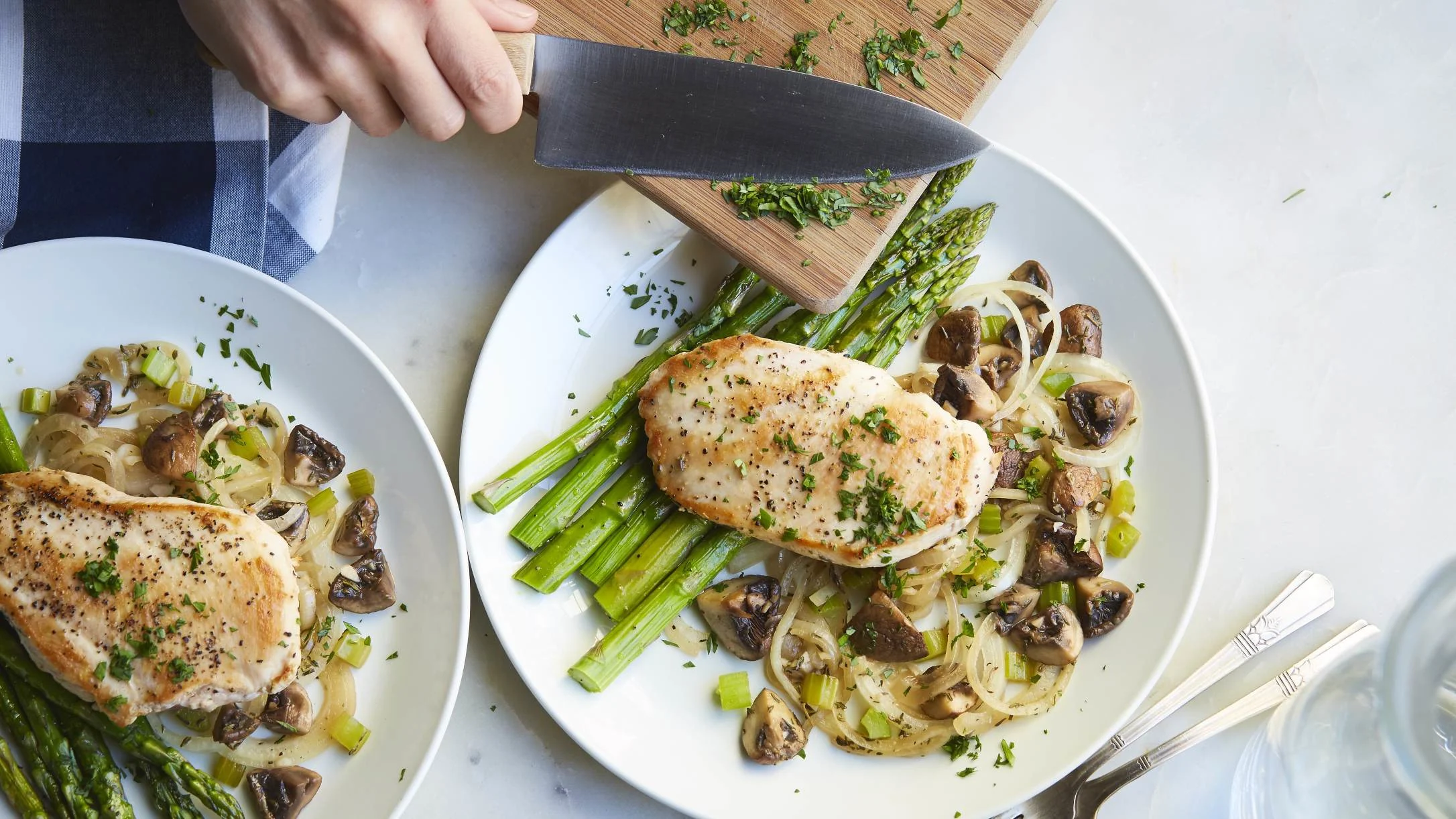 Two white plates with seared chicken breasts, asparagus, mushrooms, and onions, garnished with herbs. A hand holds a knife nearby.