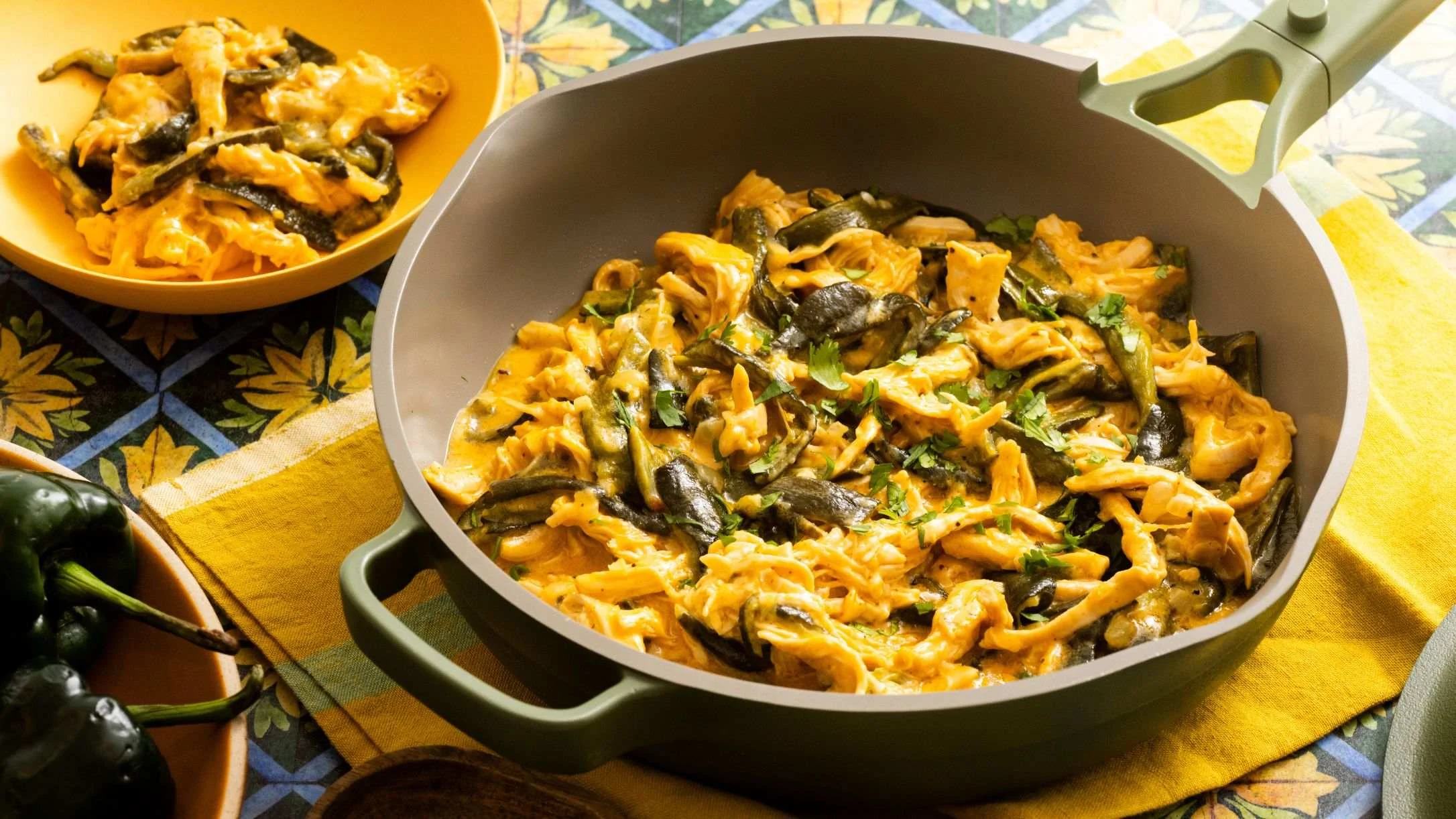 A green skillet filled with creamy shredded chicken and roasted poblano peppers, garnished with cilantro, on a yellow cloth.