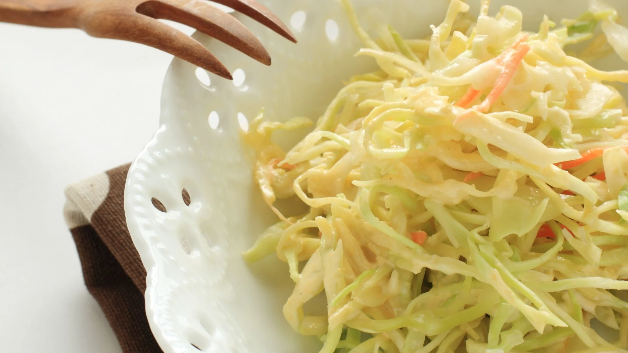 Creamy coleslaw with shredded cabbage and carrots in a white bowl, with a wooden serving fork on a brown cloth.